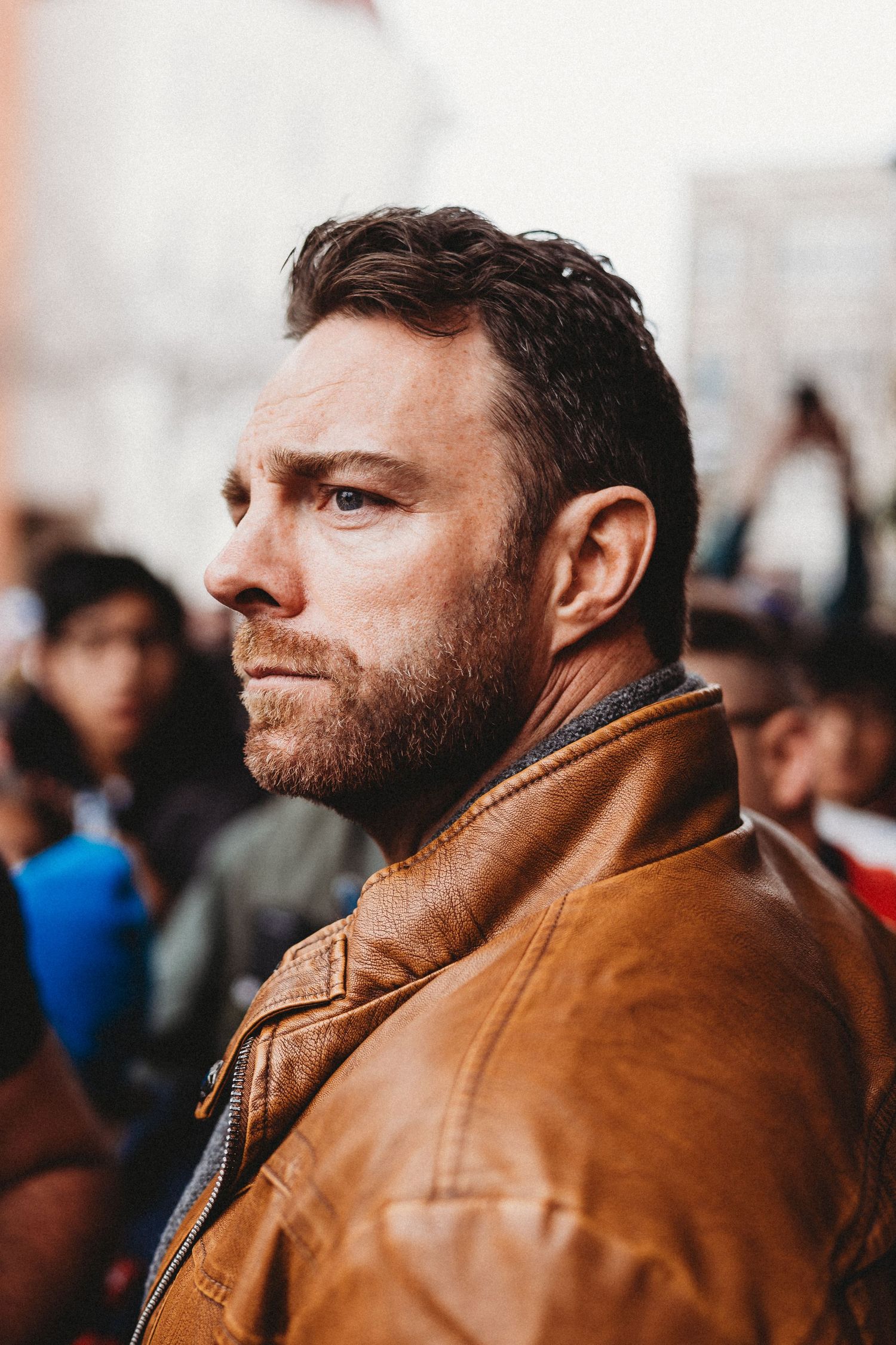 Profile view of a man with a beard wearing a brown leather jacket standing in a crowd.