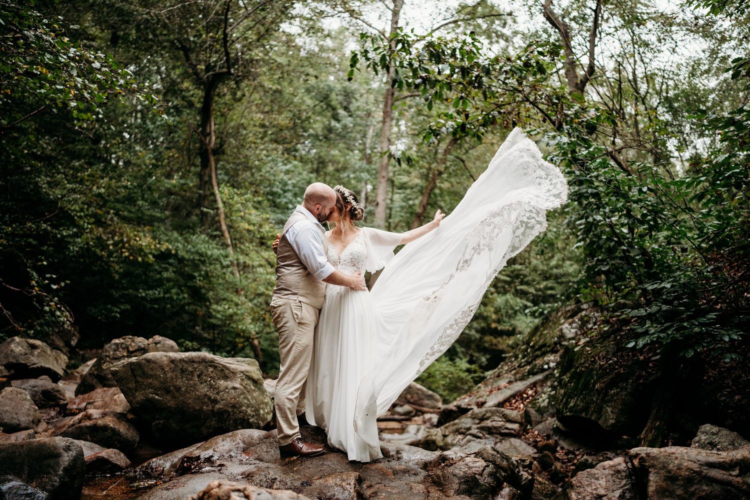 A person in a flowing white wedding dress poses gracefully on mossy rocks in a lush forest setting.
