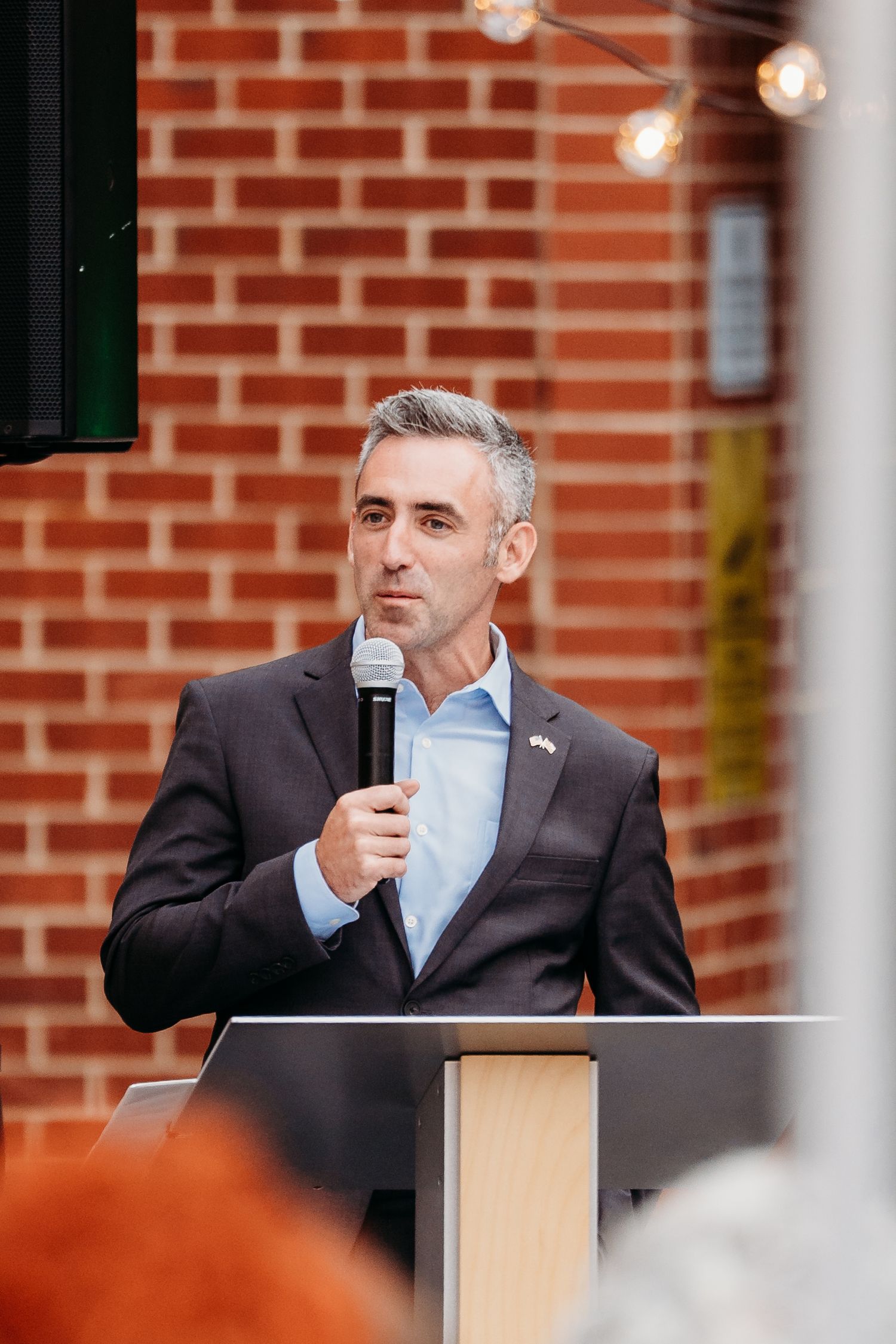 A speaker in a suit and light blue shirt addresses an audience from behind a wooden podium against a brick wall.