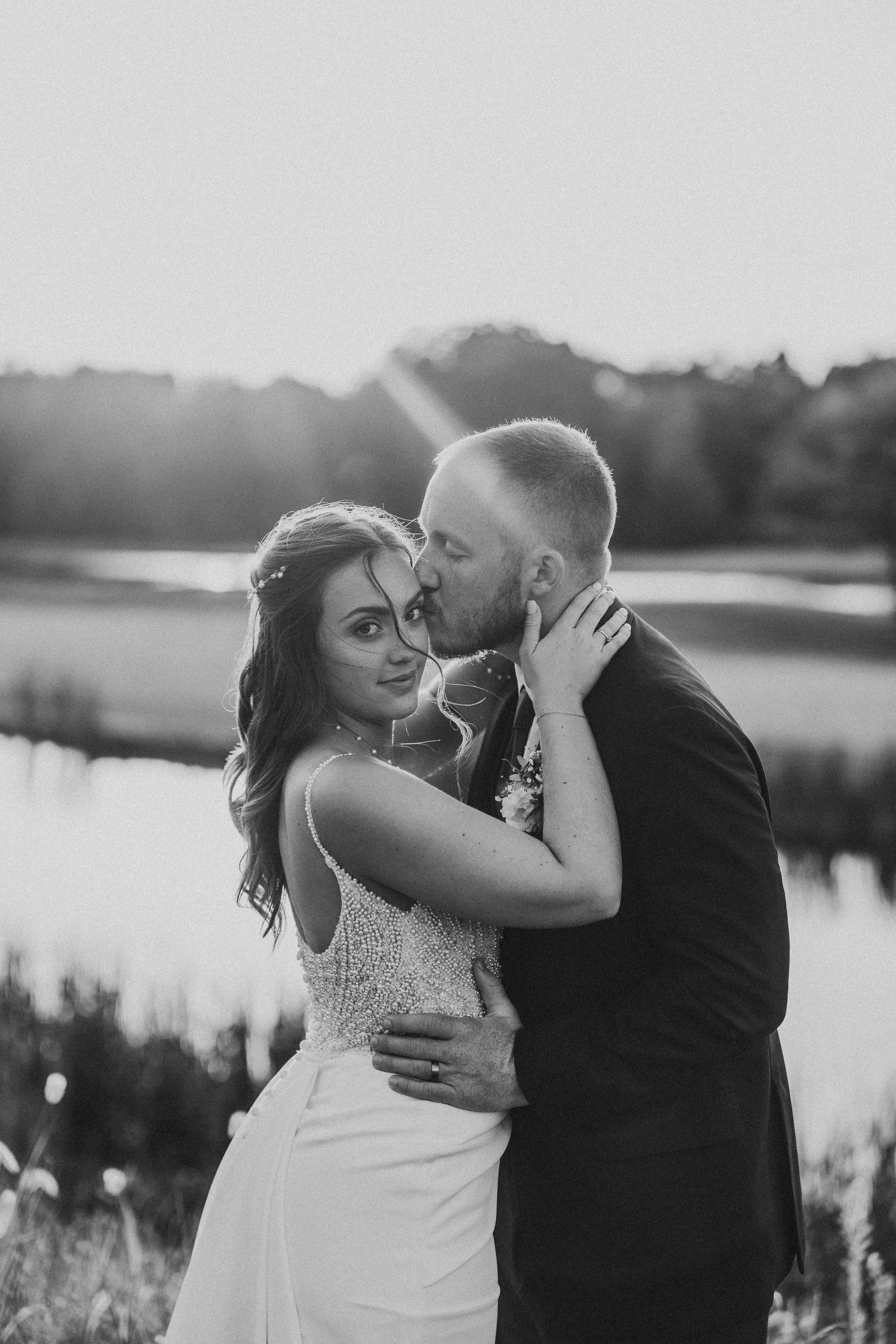 Sweet romantic moment between newlyweds by a lake during sunset, with a scenic natural backdrop.