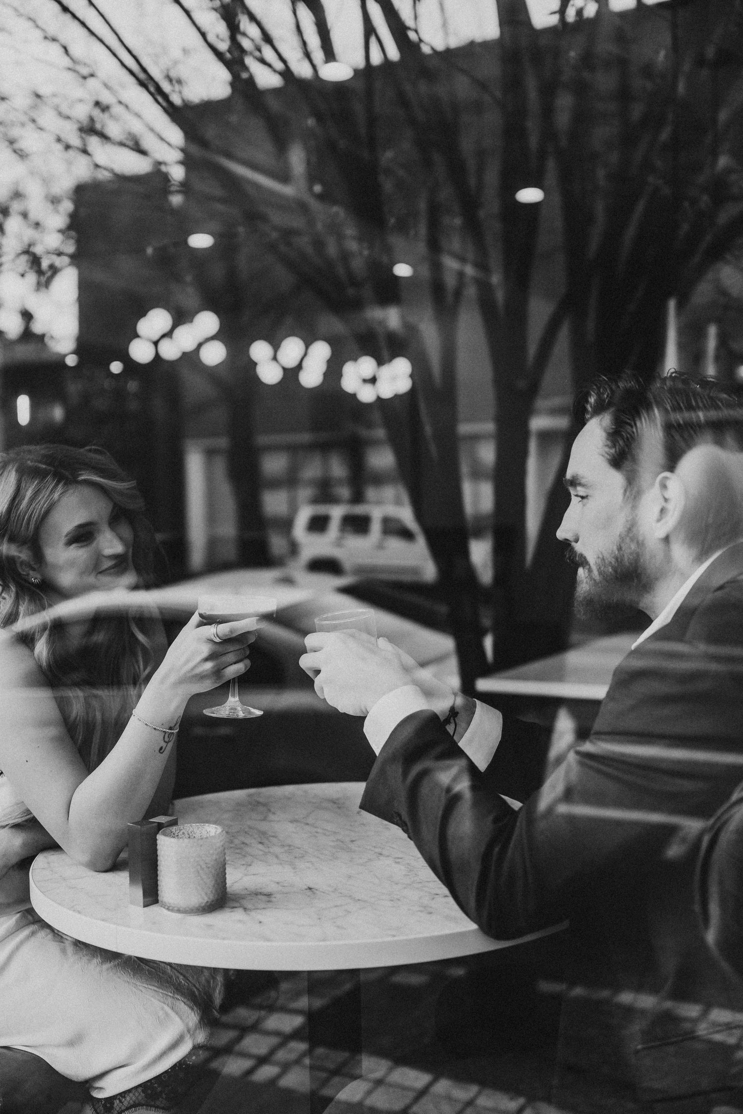 Black and white photo of people sitting at a cafe table seen through window reflections.