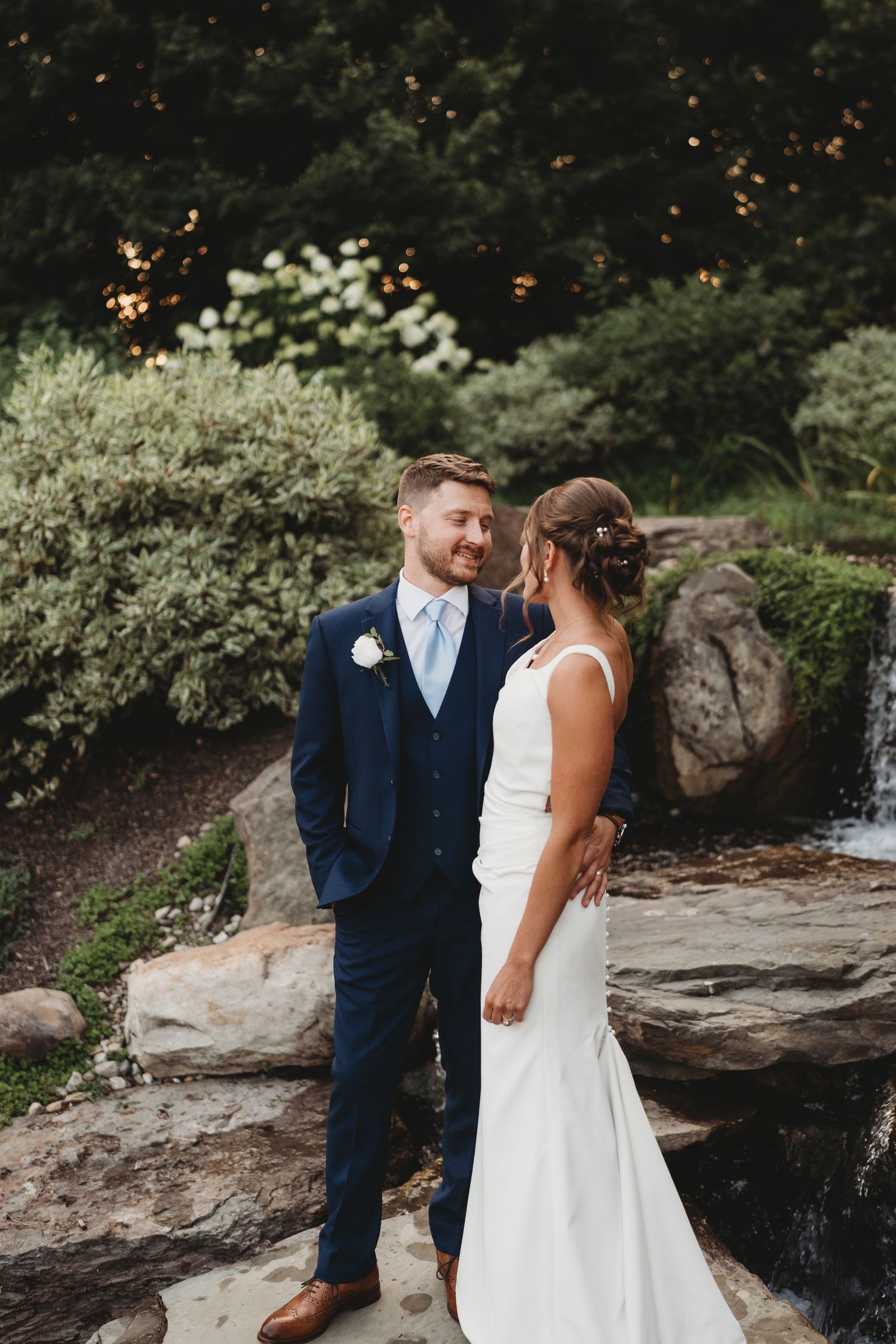 A bride and groom share a romantic moment on rocks near a waterfall, surrounded by lush greenery.