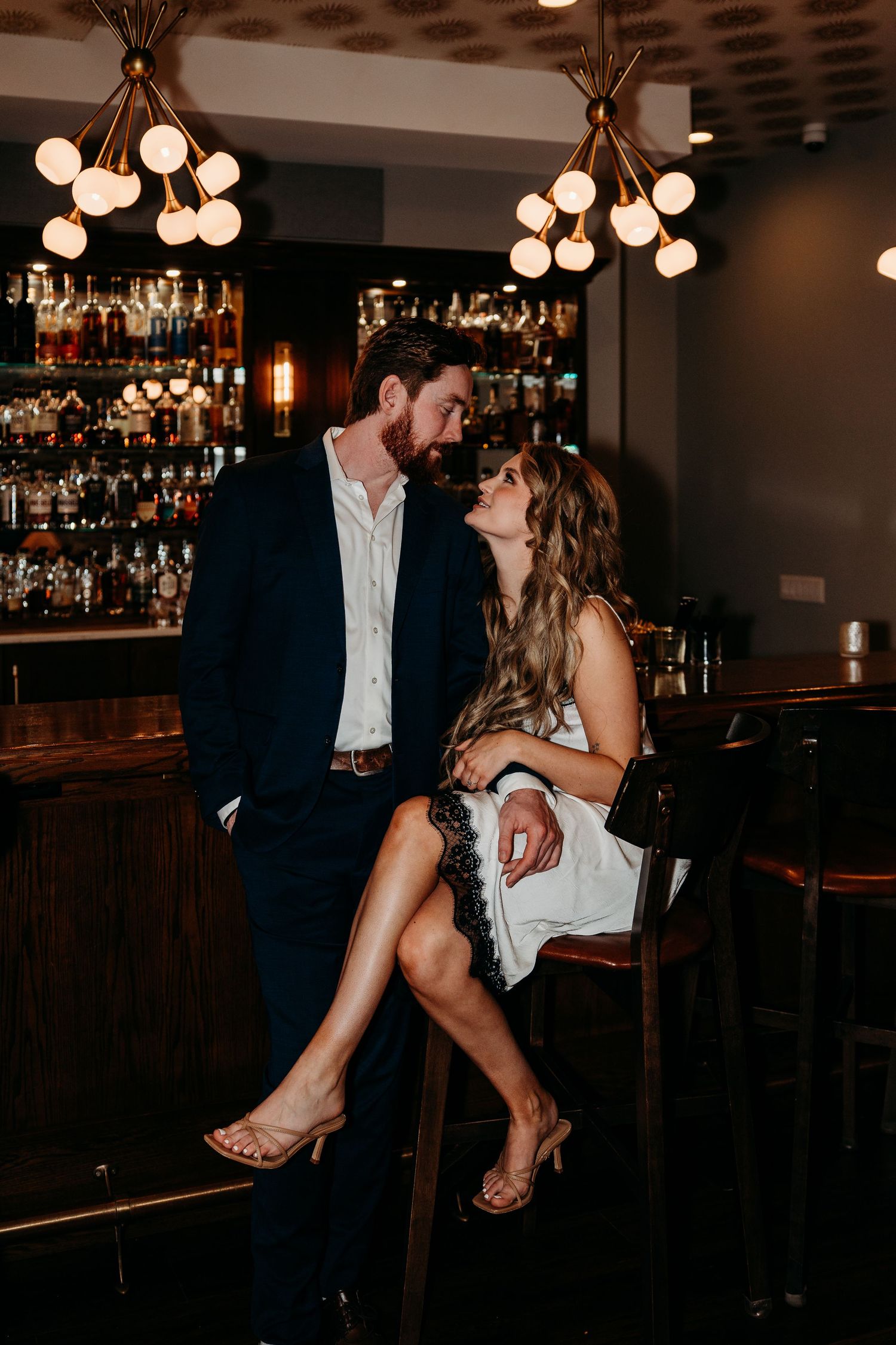 Romantic couple sharing an intimate moment at a dimly lit upscale bar with elegant globe light fixtures overhead.