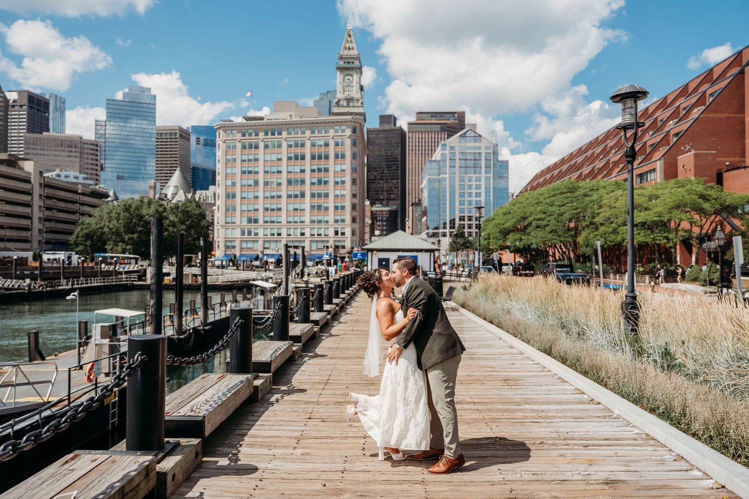 Bride and groom share a romantic kiss on a waterfront boardwalk with Boston city skyline in the background.