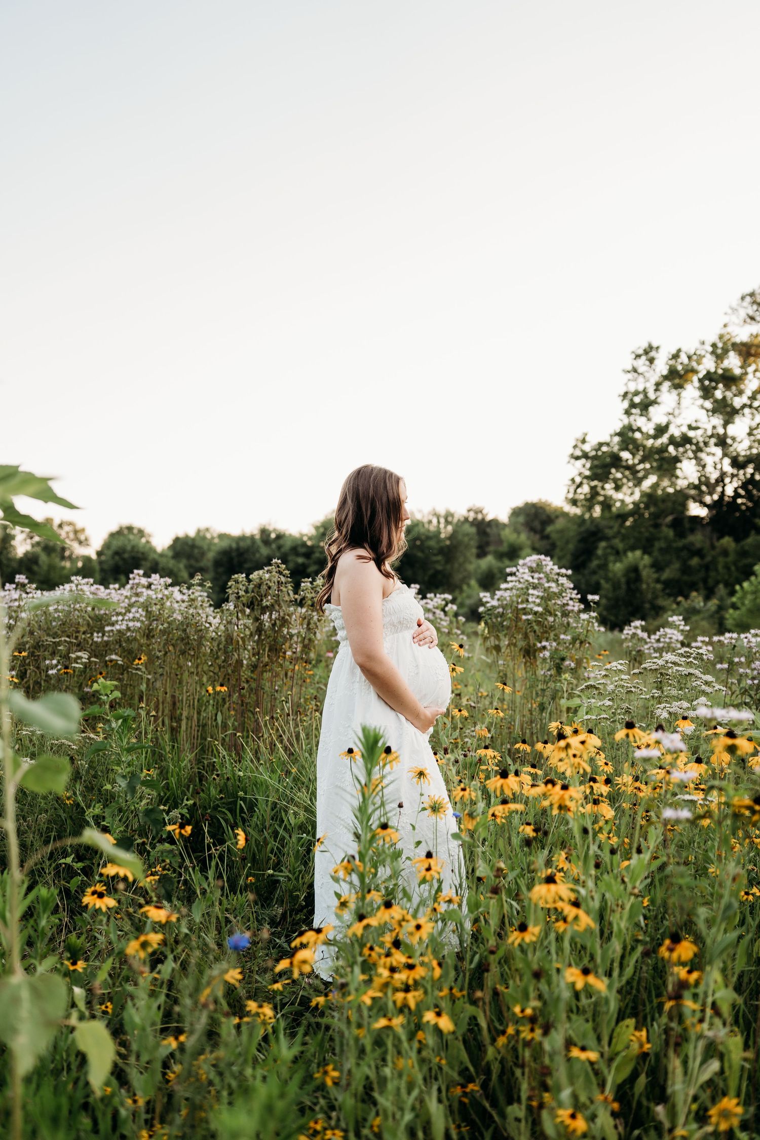Pregnant woman in white dress standing in wildflower field with yellow black-eyed Susans, cradling her baby bump outdoors.