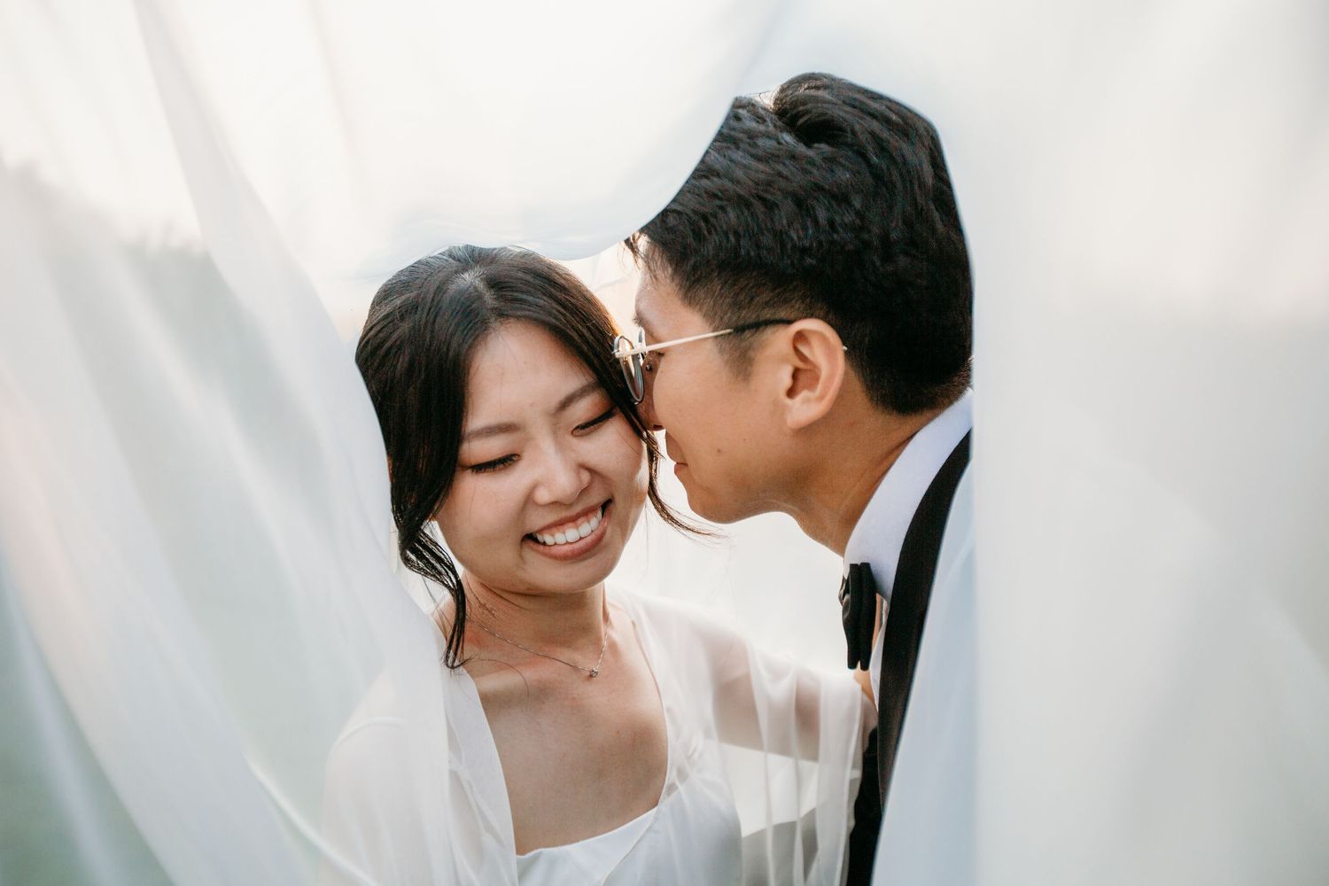 A groom kissing his bride on the cheek as she smiles joyfully, framed by a flowing white veil.