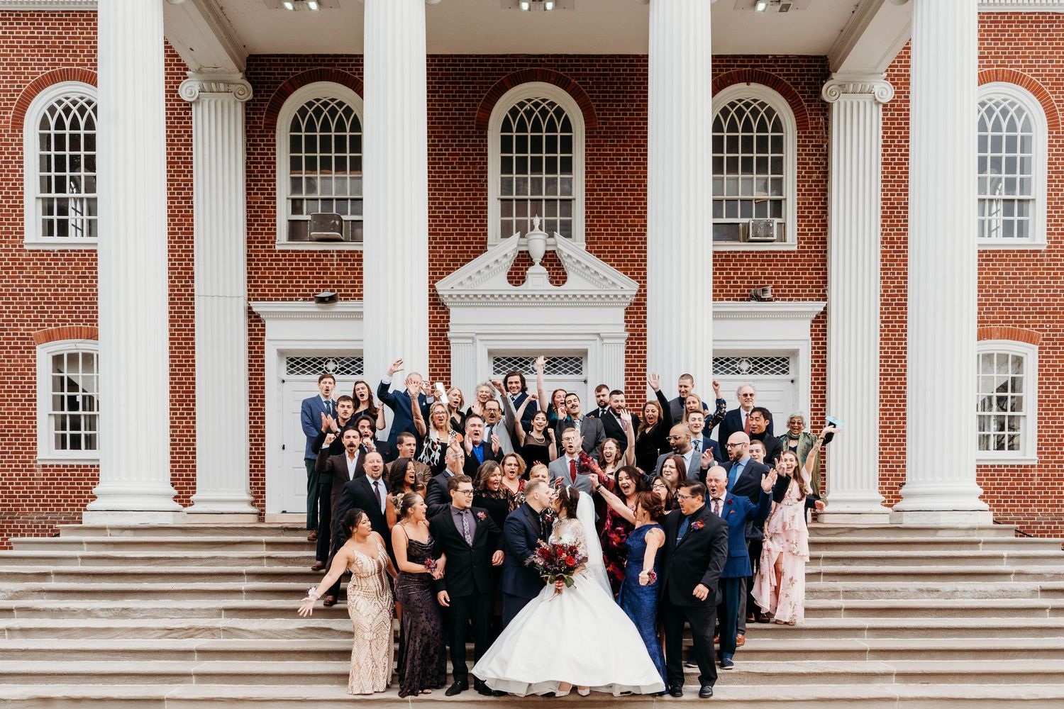 A large wedding party poses on the steps of a grand brick building with white columns and arched windows.