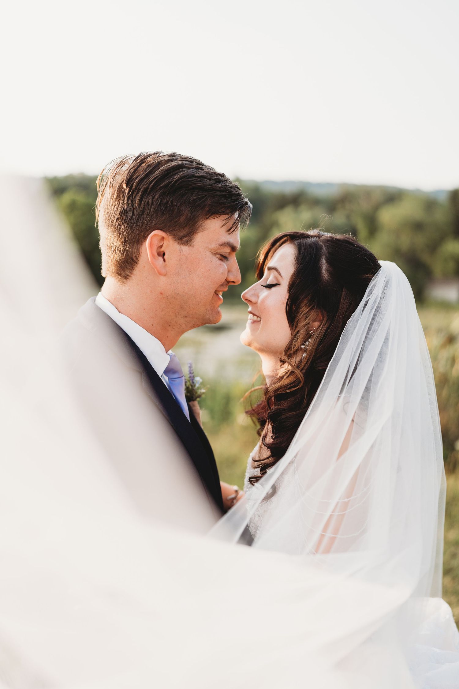 A romantic wedding portrait with a bride in white veil and groom sharing an intimate moment outdoors in natural lighting.