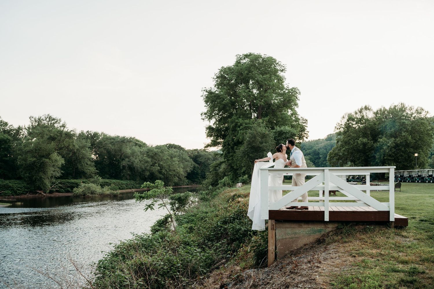 Couple standing on a white bridge over a river surrounded by lush green trees at dusk.