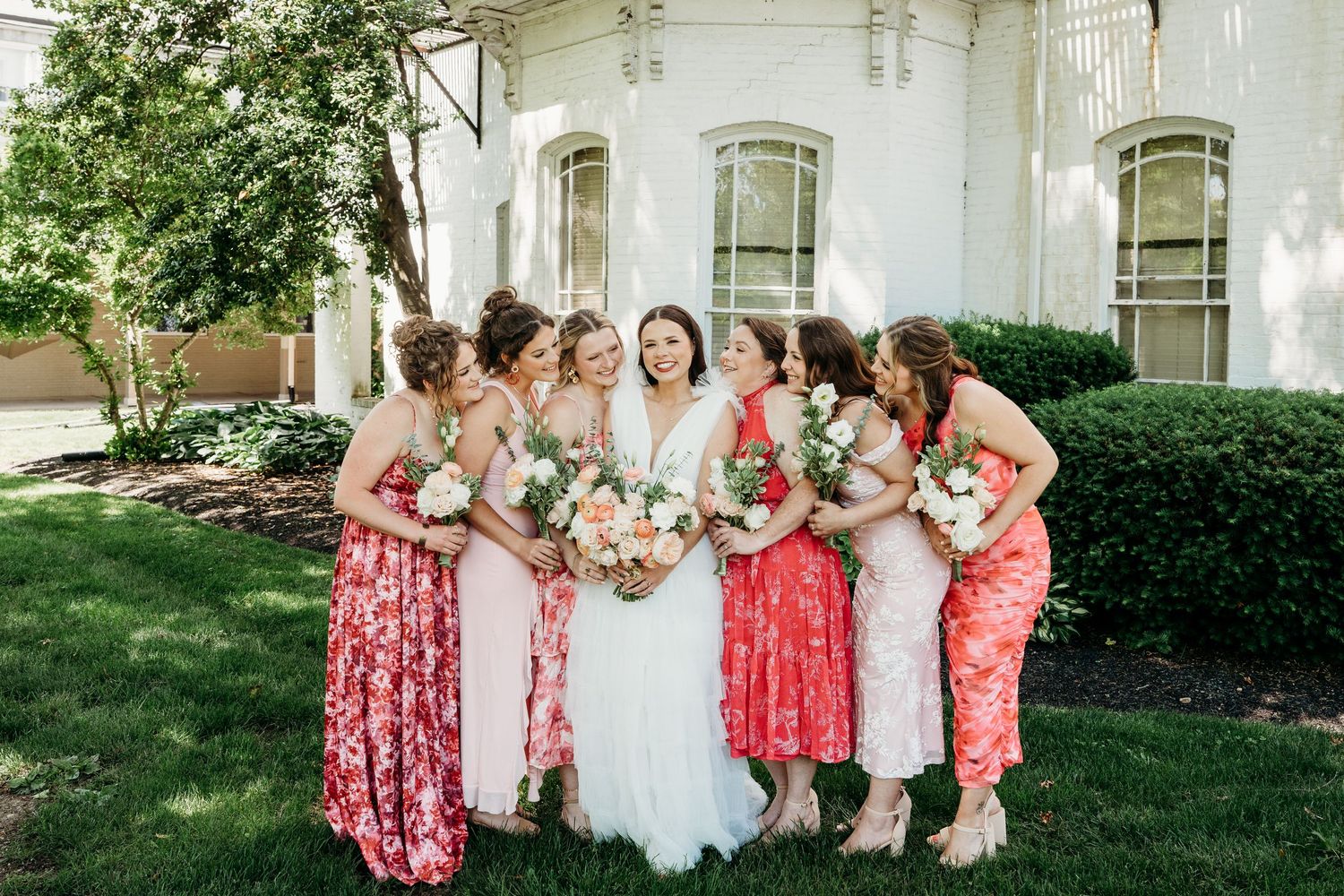 Bride in white gown with bridesmaids in coral floral dresses holding bouquets outside elegant white building.