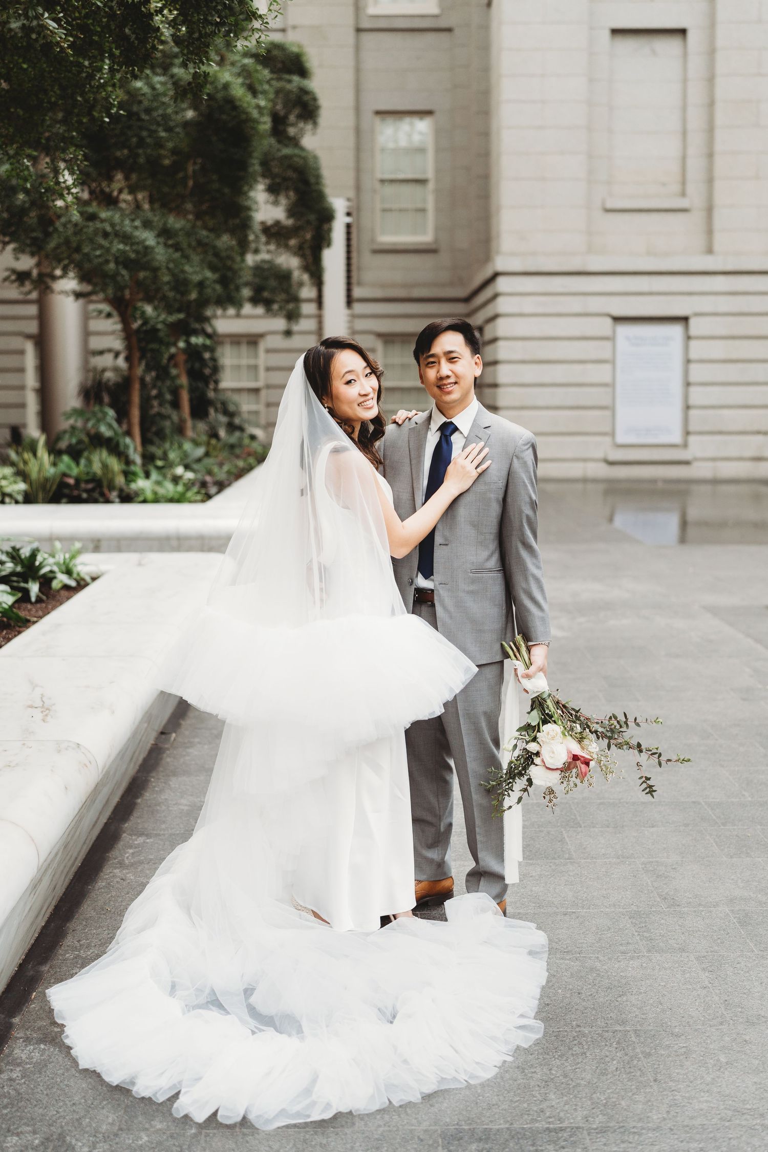 Newlyweds pose outdoors in formal attire with flowing white wedding gown against classic architecture.