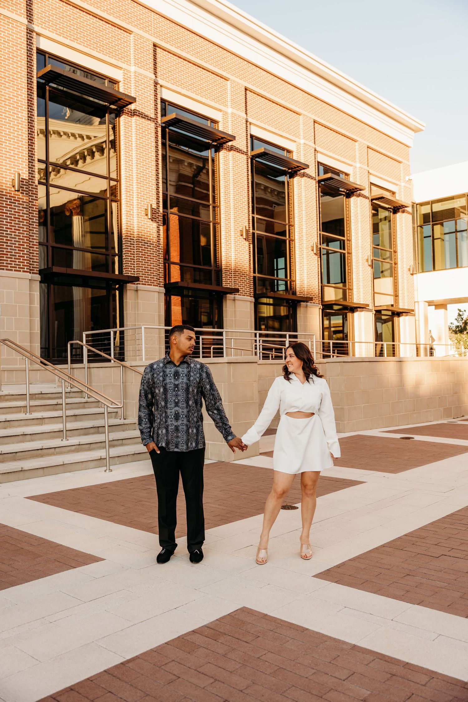 A couple holding hands while walking outside a modern brick building during golden hour.