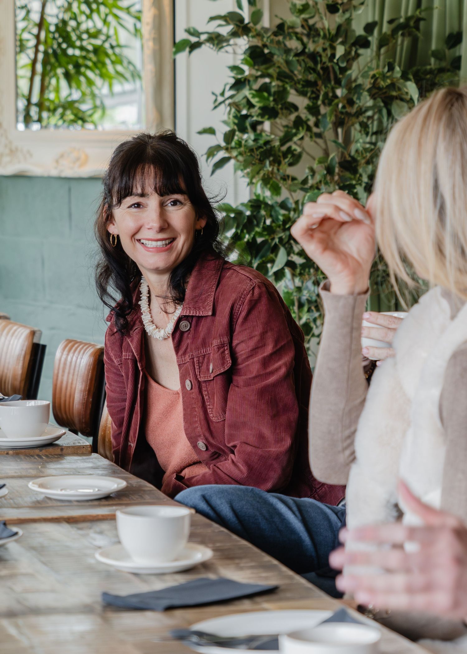 Two women smiling and chatting at a rustic cafe table with coffee cups, surrounded by lush green plants.