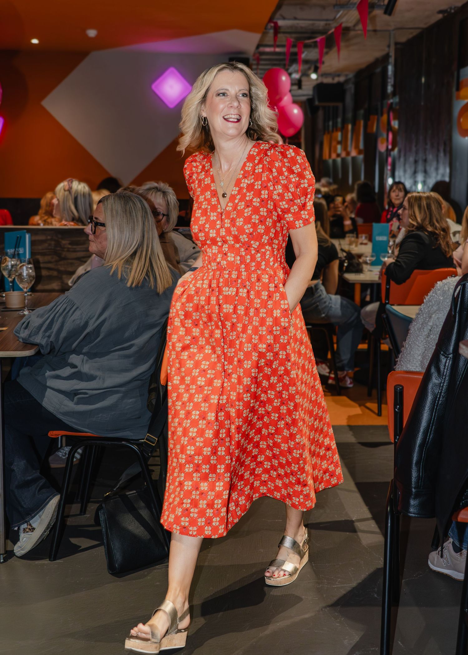 Woman in orange patterned midi dress with gold sandals posing in a vibrant restaurant setting.