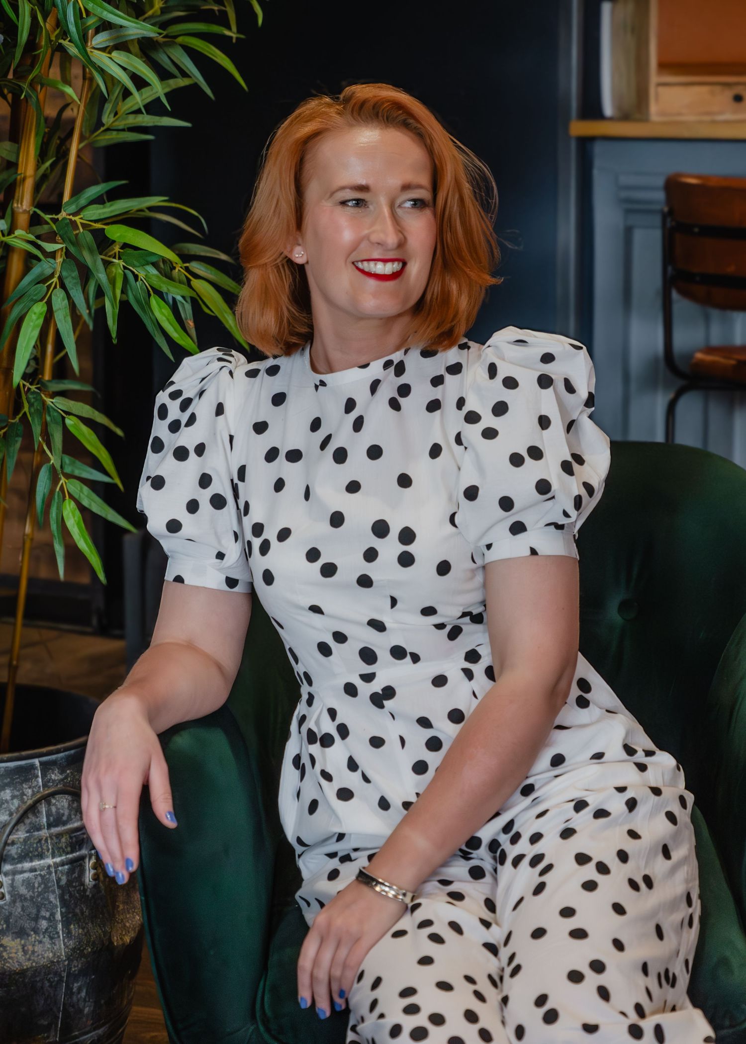 Woman in white polka dot outfit sitting on green velvet chair, smiling with red hair and red lipstick.