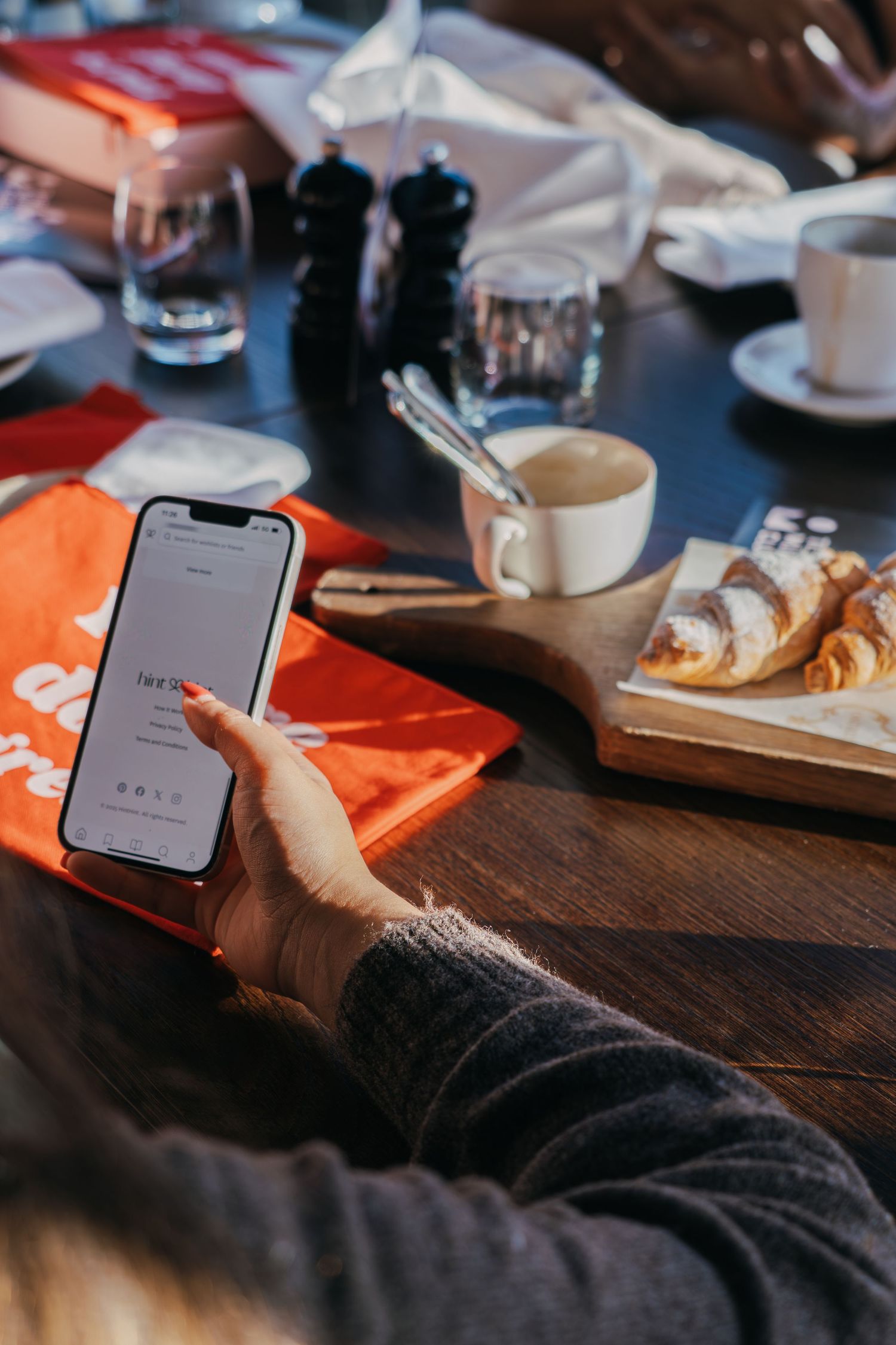 Restaurant table setting with phone, pastries, coffee cup and black salt shakers on wooden surface.