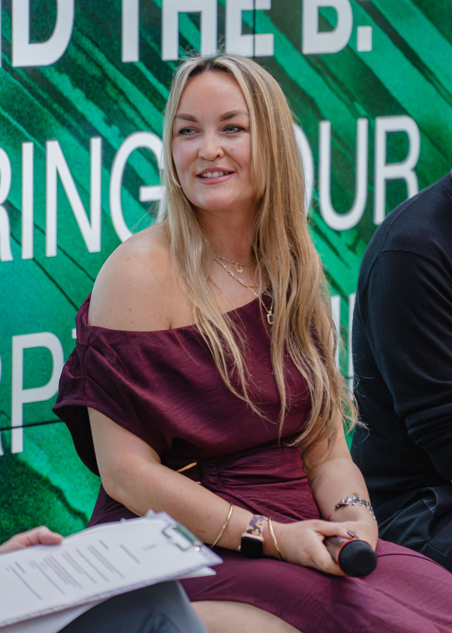 A person in a burgundy dress sits at a table against a bright green backdrop with text.