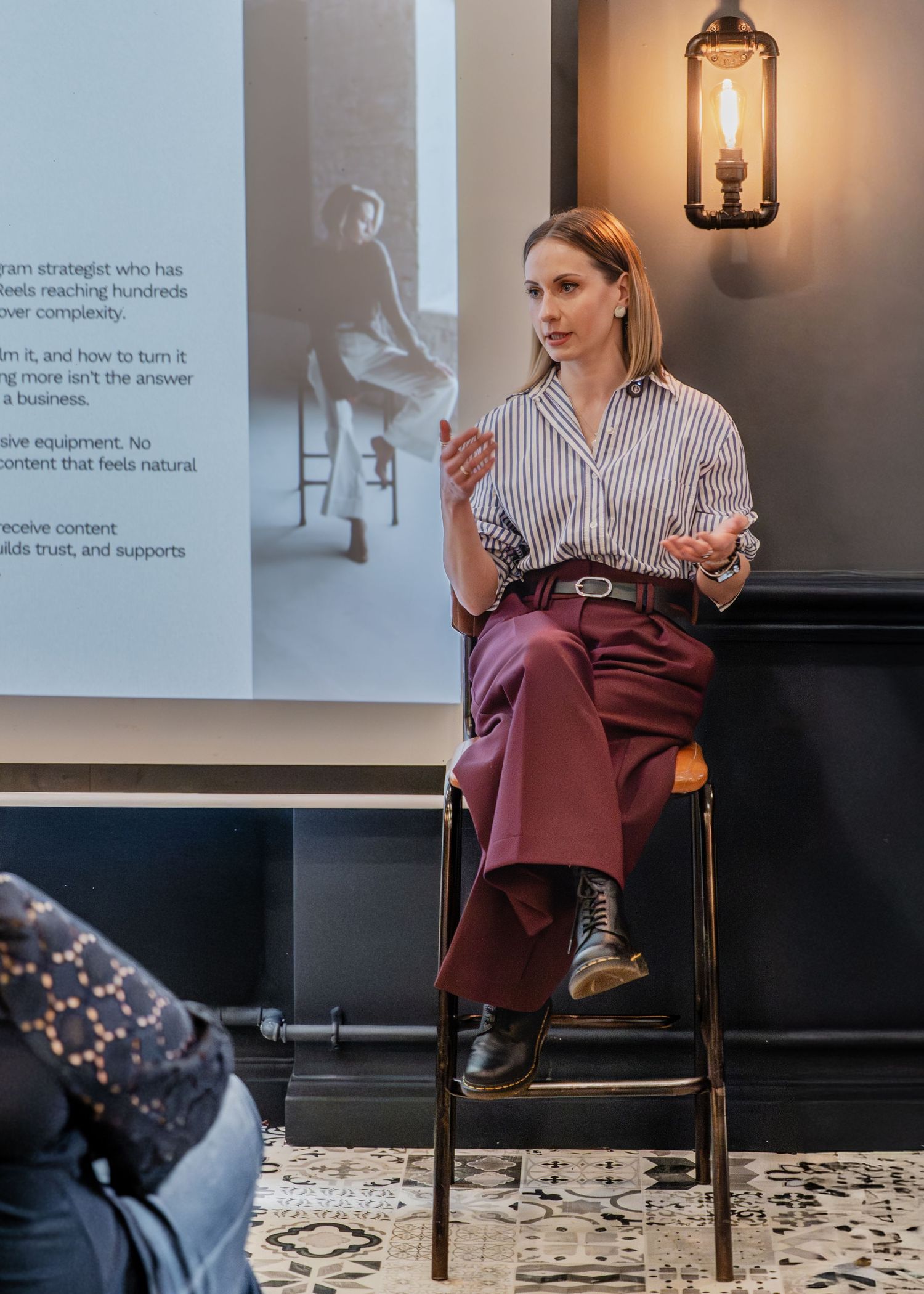 Woman in striped shirt and burgundy trousers sits on stool presenting at a business seminar with projection screen behind her.