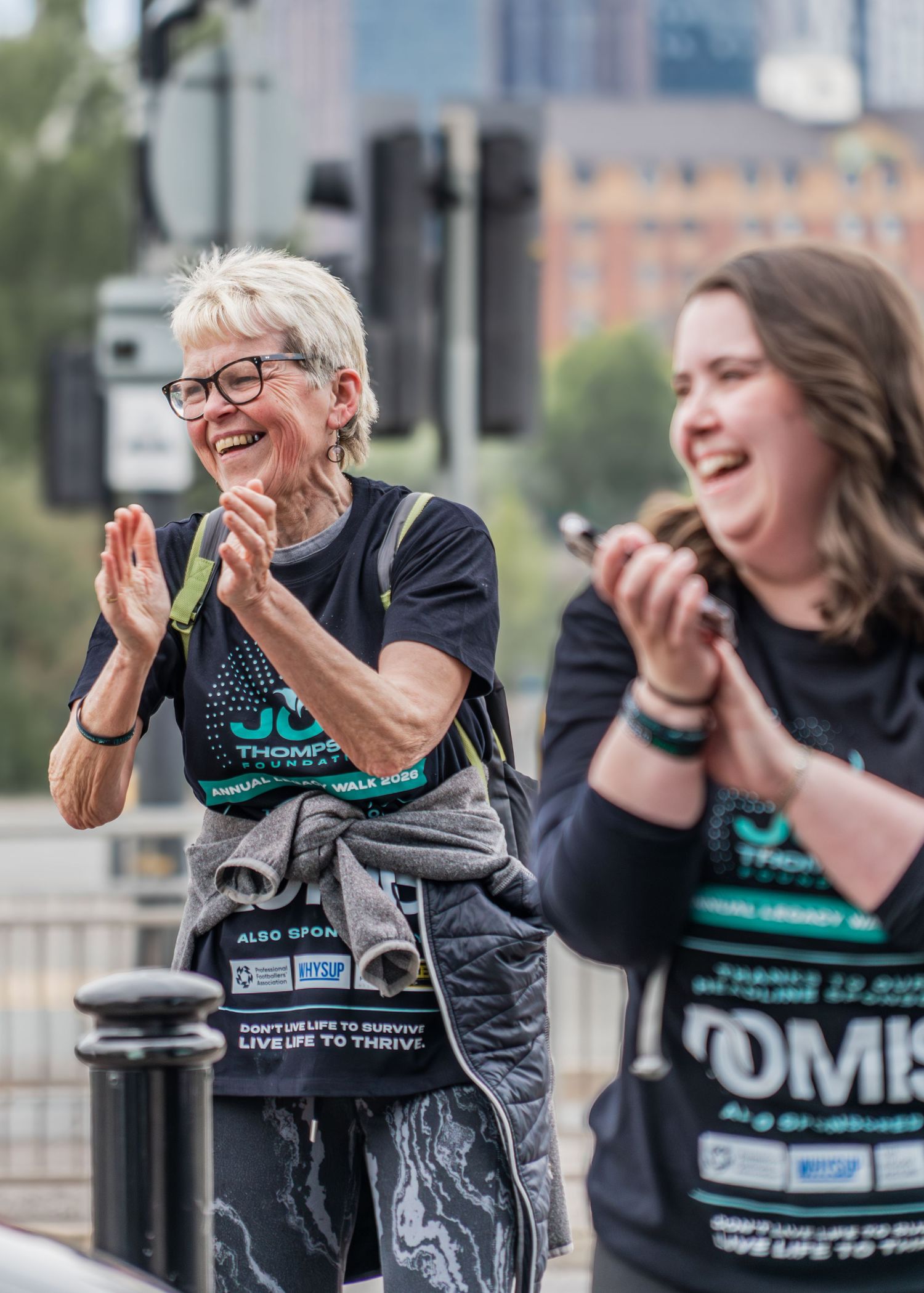 Two women in charity walk t-shirts laughing and clapping outdoors at a fundraising event.