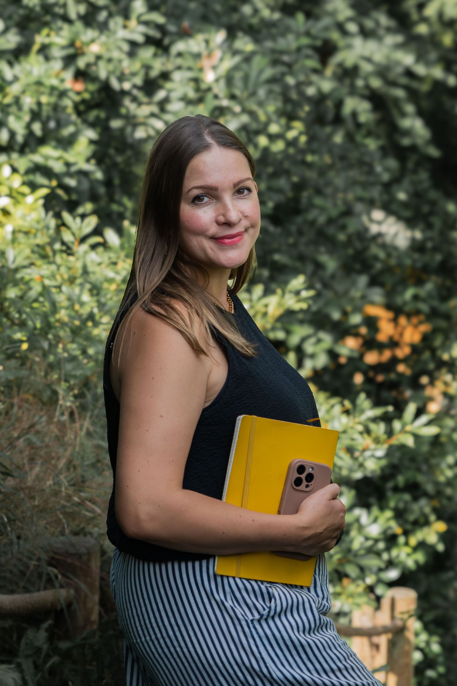 A person in a black top and striped skirt holds a yellow notebook while posing outdoors among green foliage.