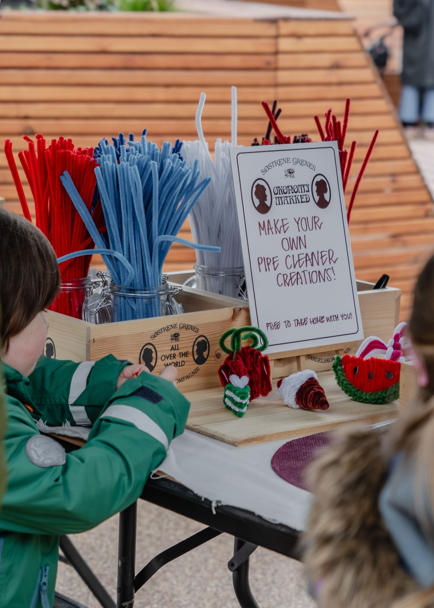 Hands in green sleeves reach for colorful pipe cleaners and craft supplies arranged in labeled containers at a crafting station.
