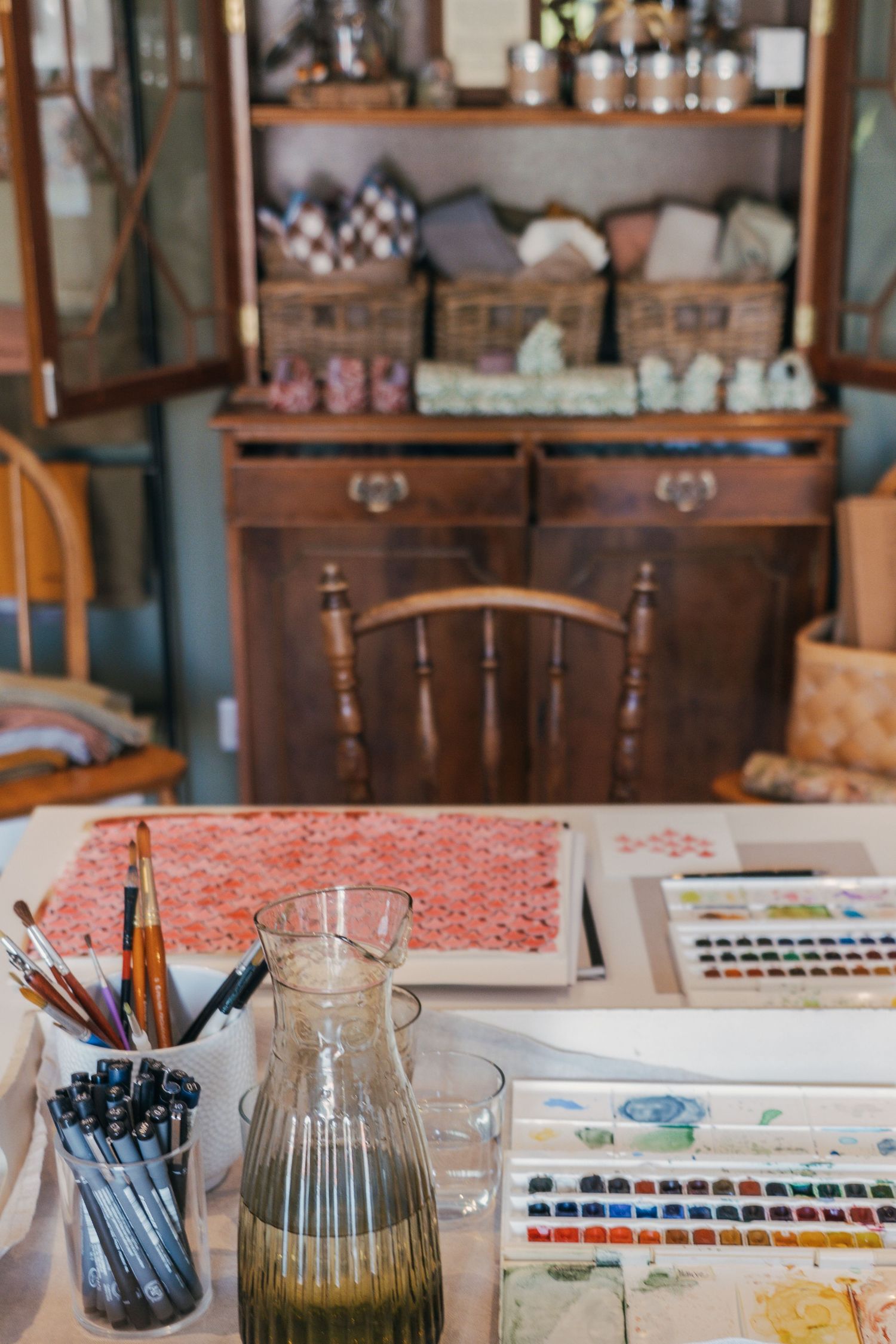 Art supplies and watercolor palettes laid out on a table with vintage furniture and glass bottles in the background.