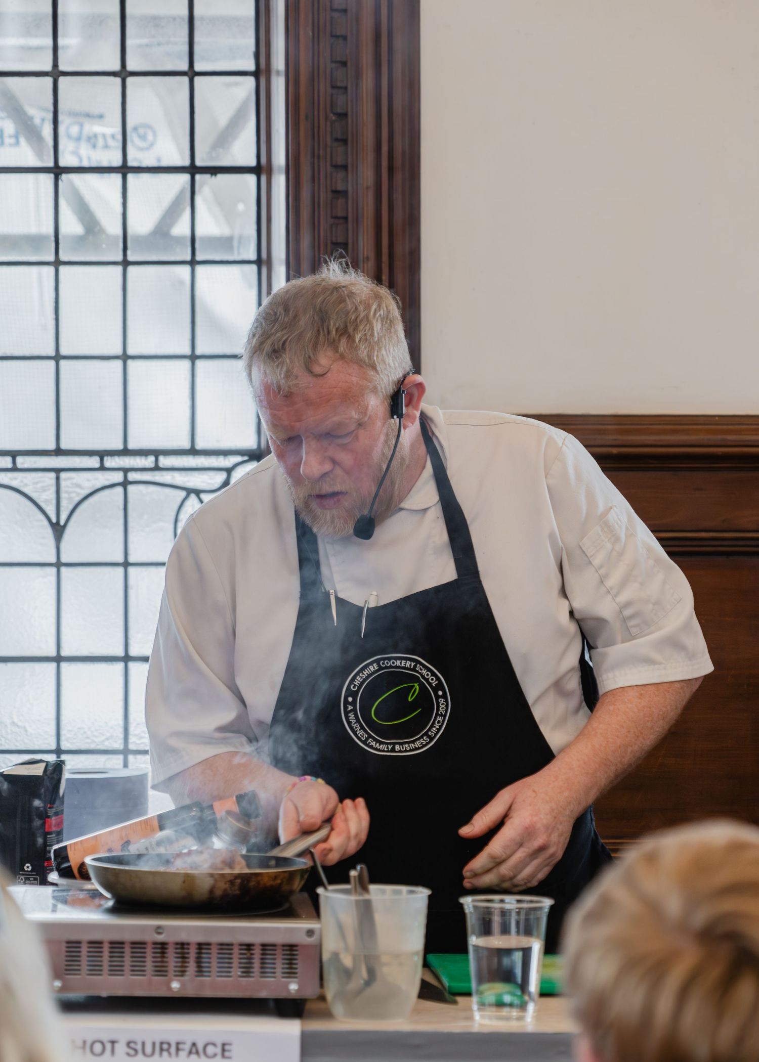 A chef in a black apron demonstrates cooking techniques at a culinary demonstration station while steam rises from the pan.