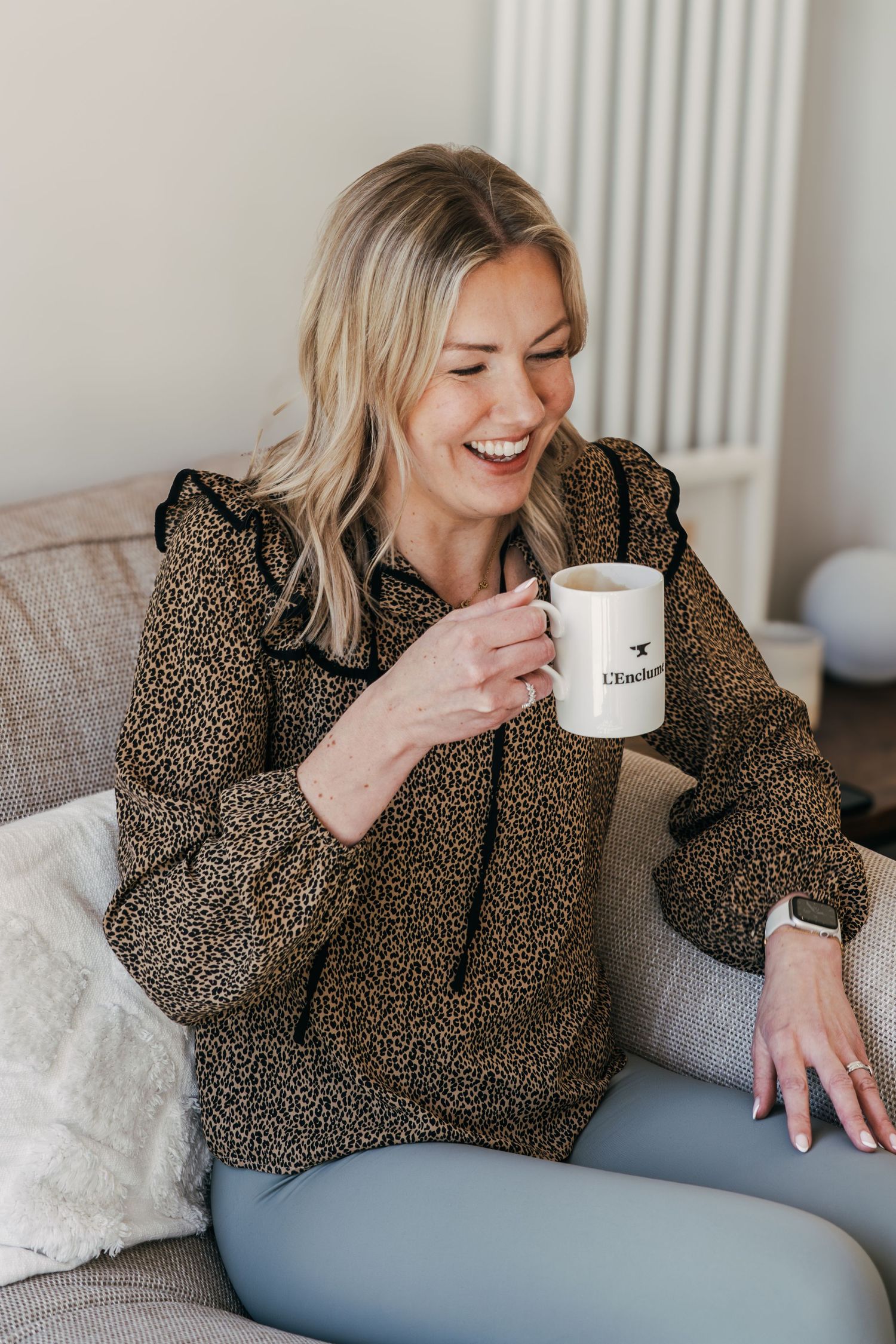 A person in a leopard print top sits on a blue couch while laughing and holding a white coffee mug.