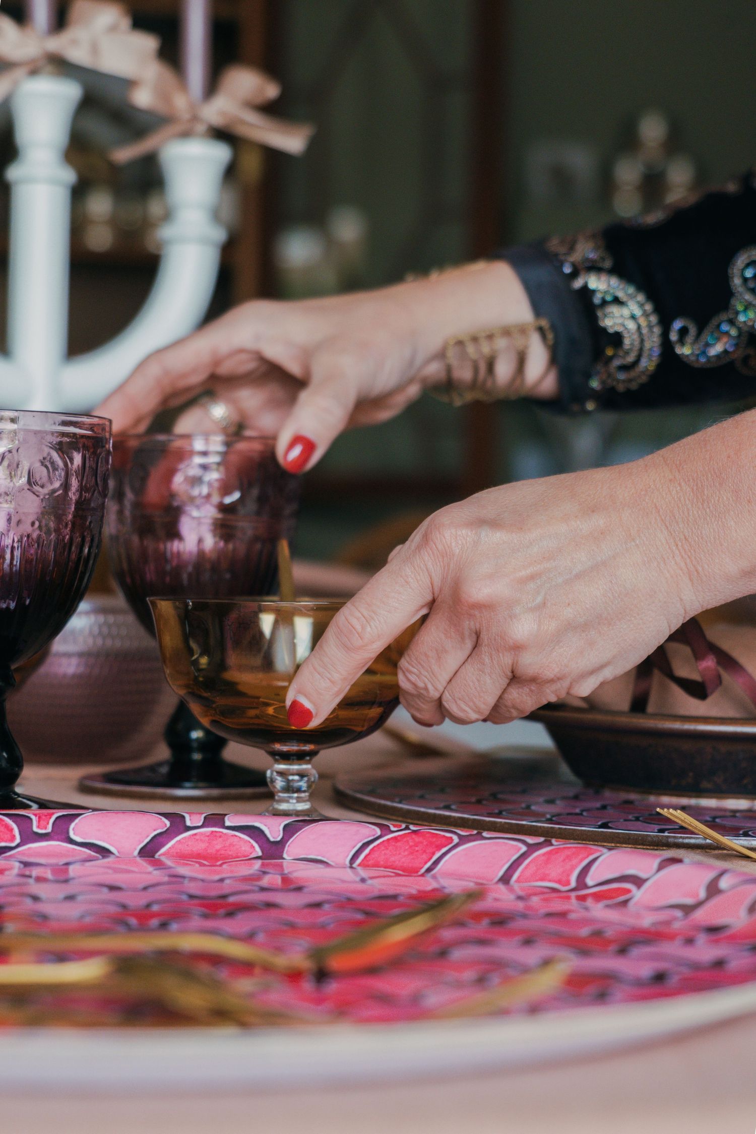 Hands with red nail polish pour wine into vintage purple glasses on a decorated pink table setting.