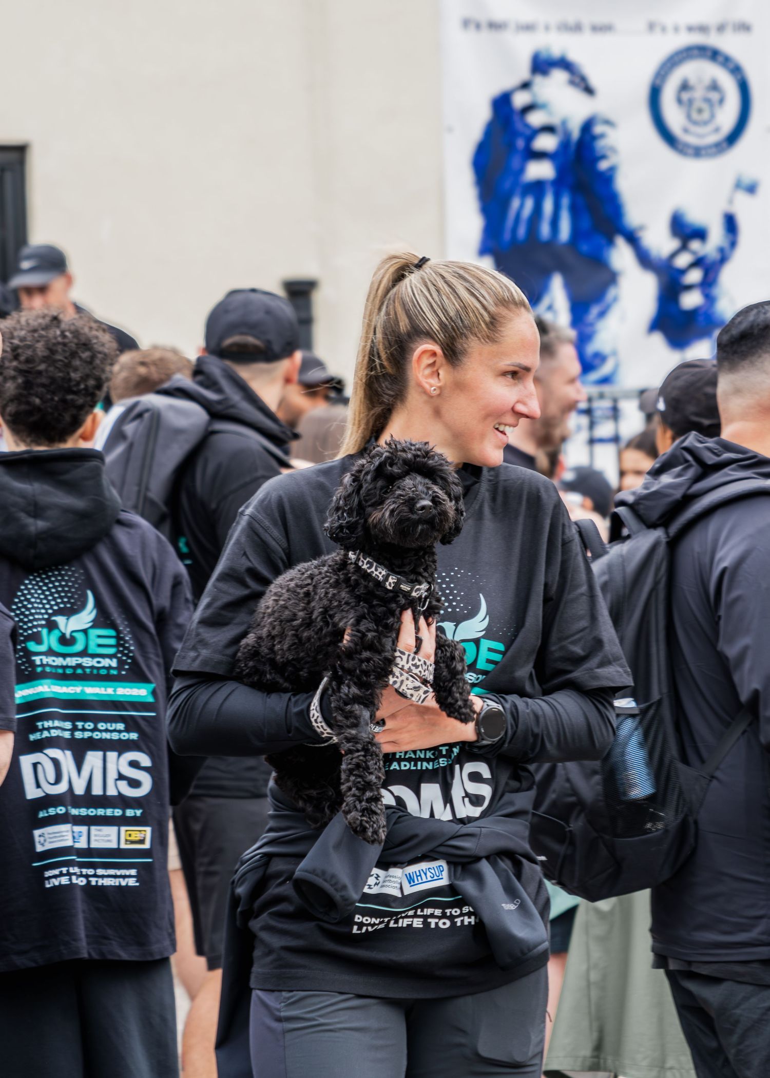Smiling woman holds a black curly dog at a Joe Thompson charity fundraising event outdoors.