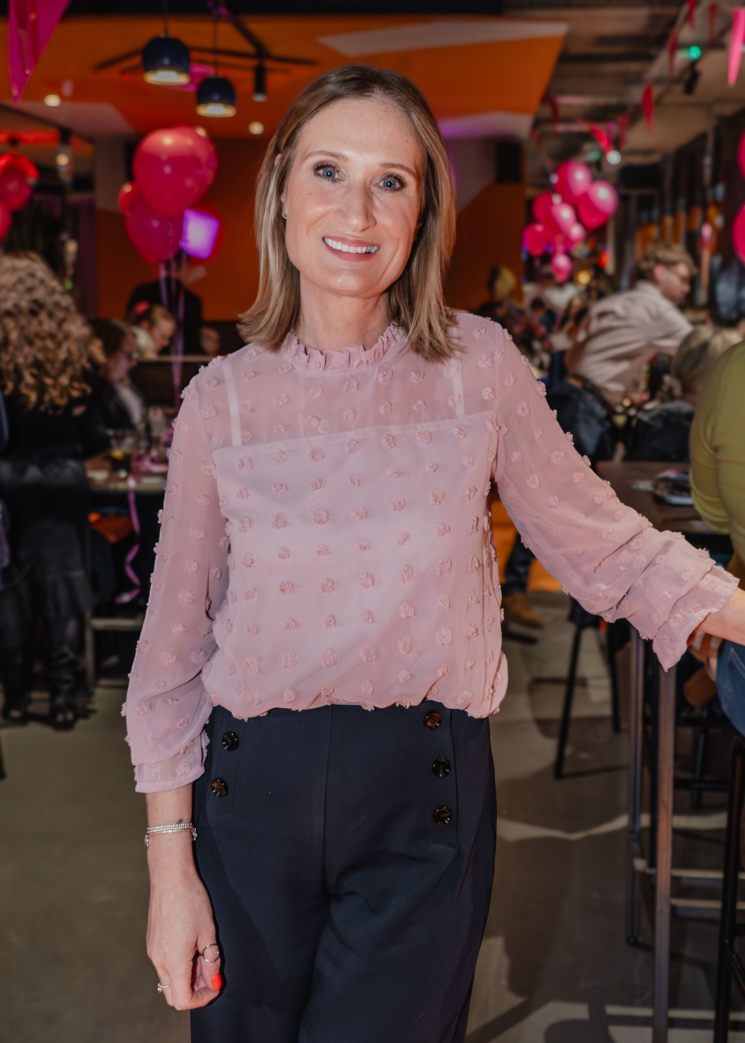 Woman in pink textured blouse and navy trousers smiling at a festive indoor event with pink balloons.