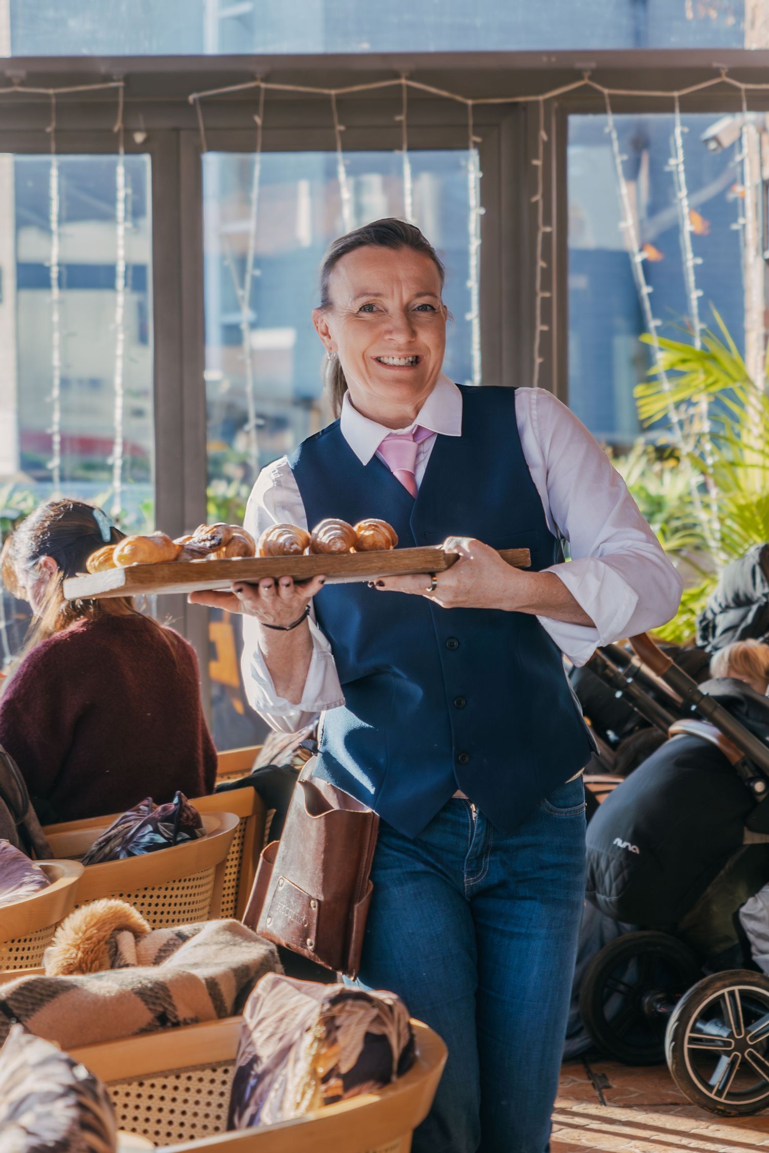 Restaurant server carrying multiple plates of food along a rattan chair lined dining room with window views.