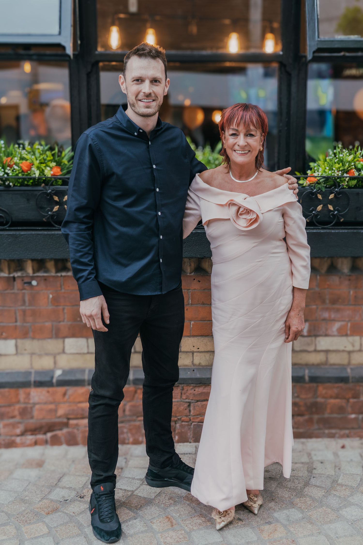 A couple stands together in formal attire outside a brick building with window planters.