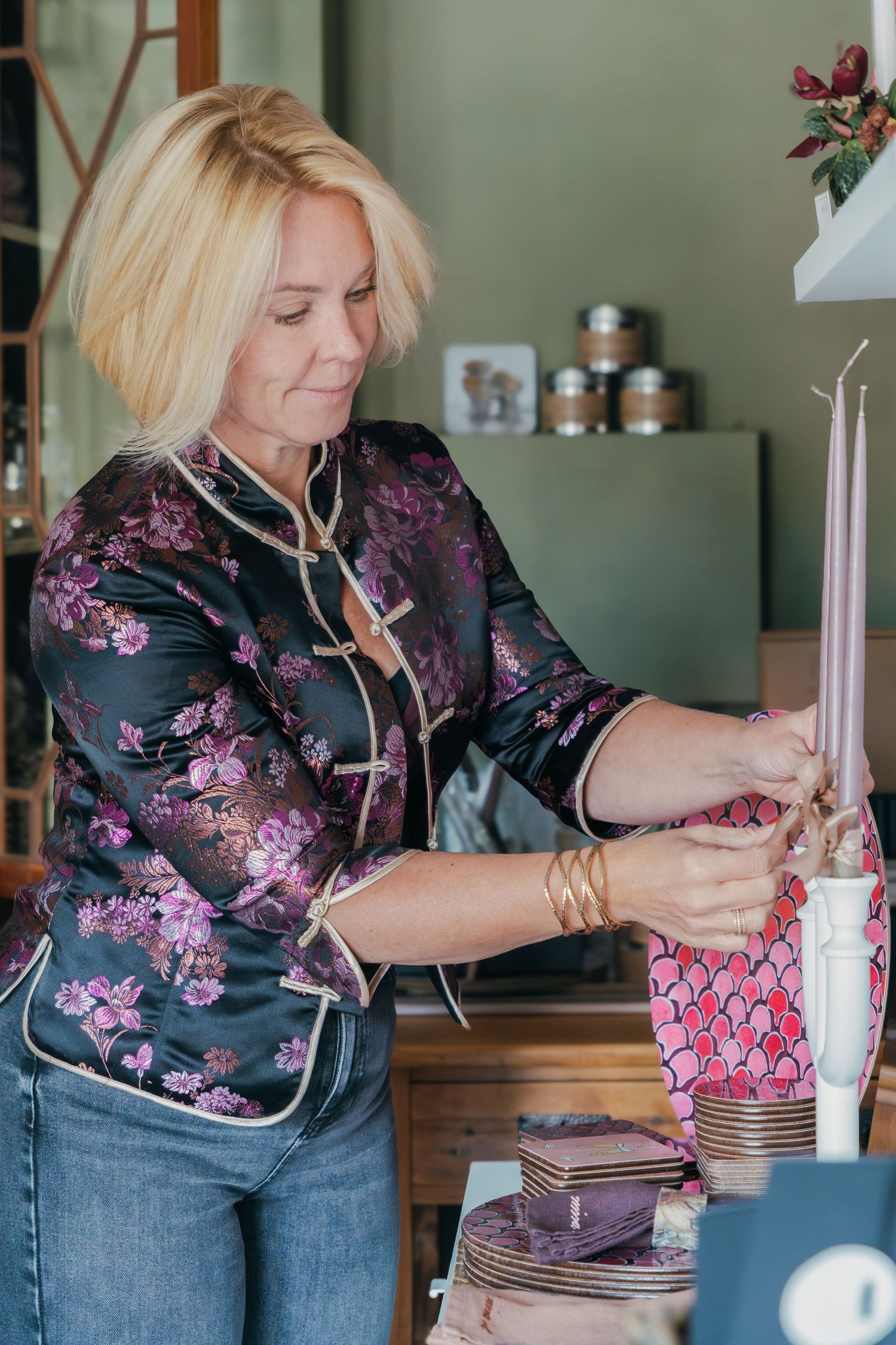 A person in a floral print top crafts with candles and decorative materials at a wooden table.
