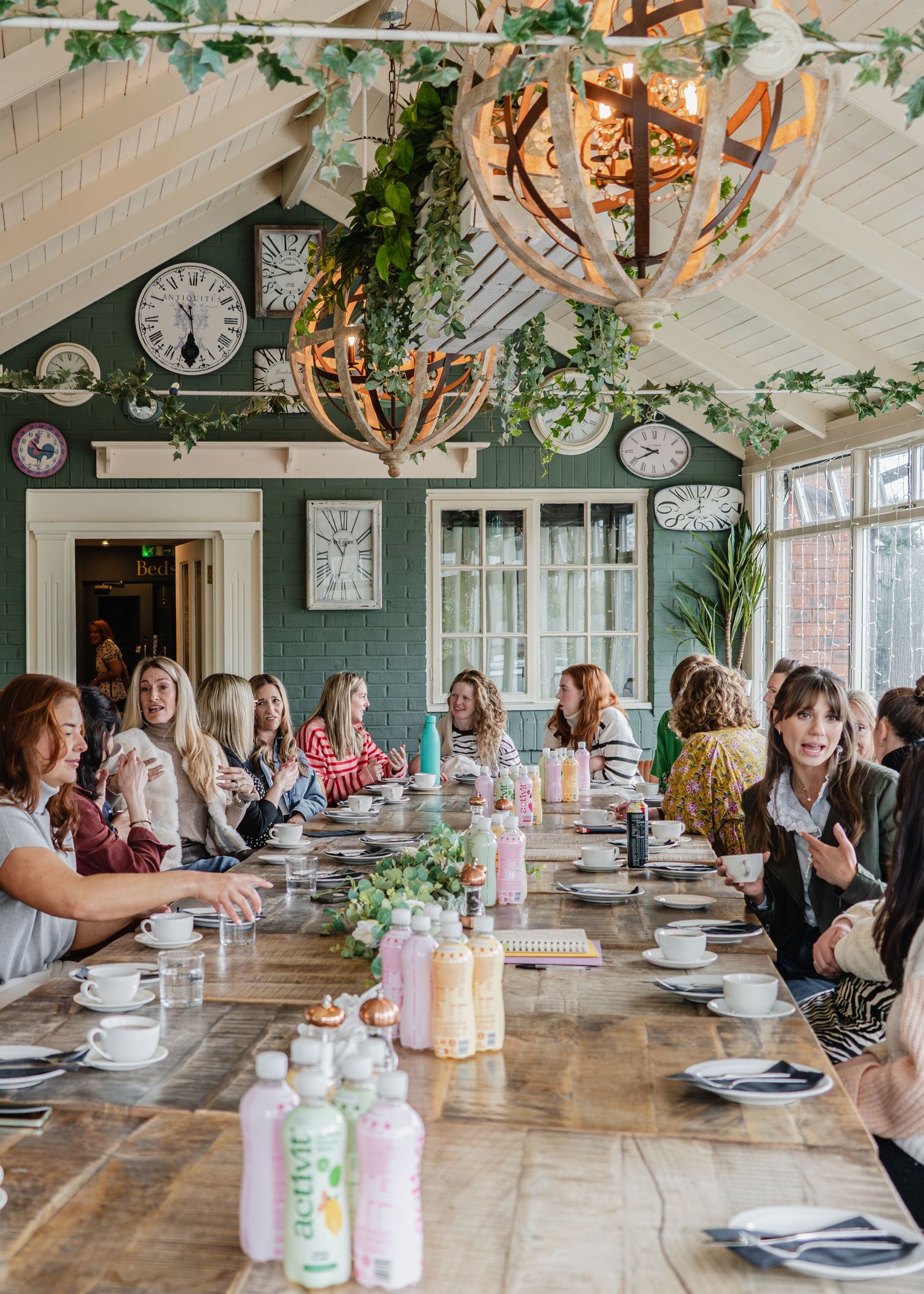 Women gathered around a long wooden table for a group luncheon in a charming green-walled restaurant with clock decor.