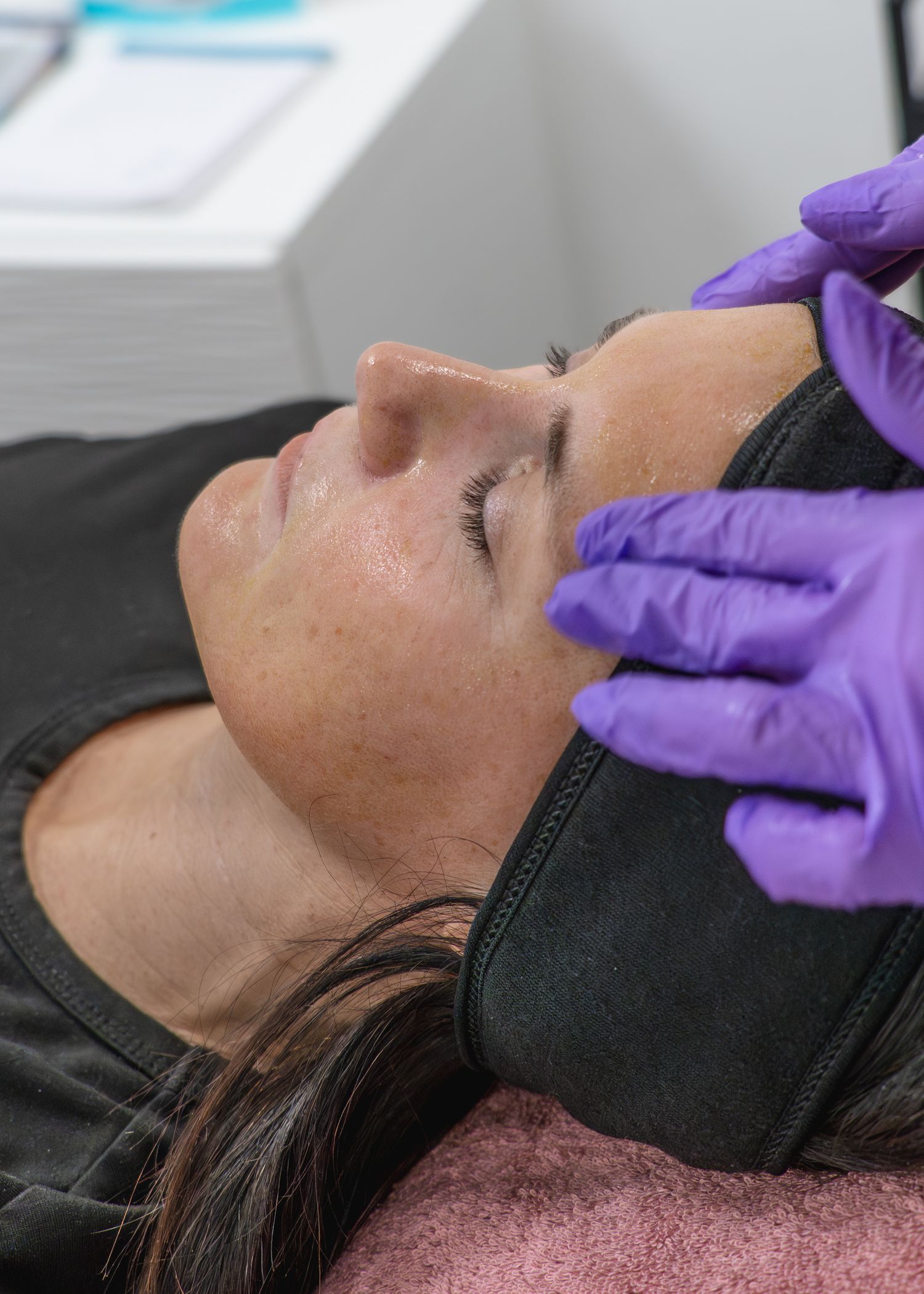 A woman receiving a facial treatment from a practitioner wearing purple gloves in a clinical setting.