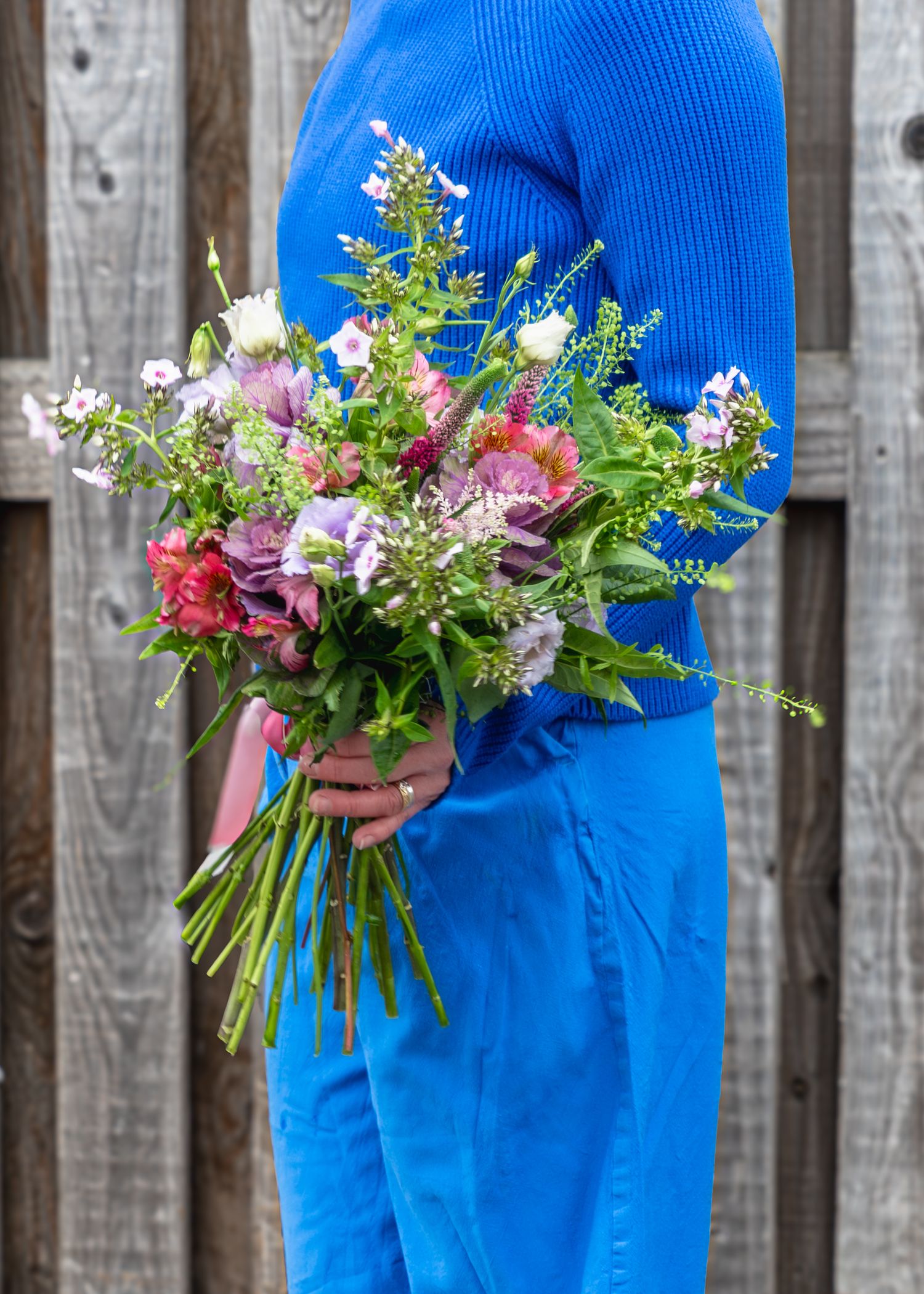 Three vibrant flower bouquets held against a blue dress with wooden fence backdrop create a colorful spring display.