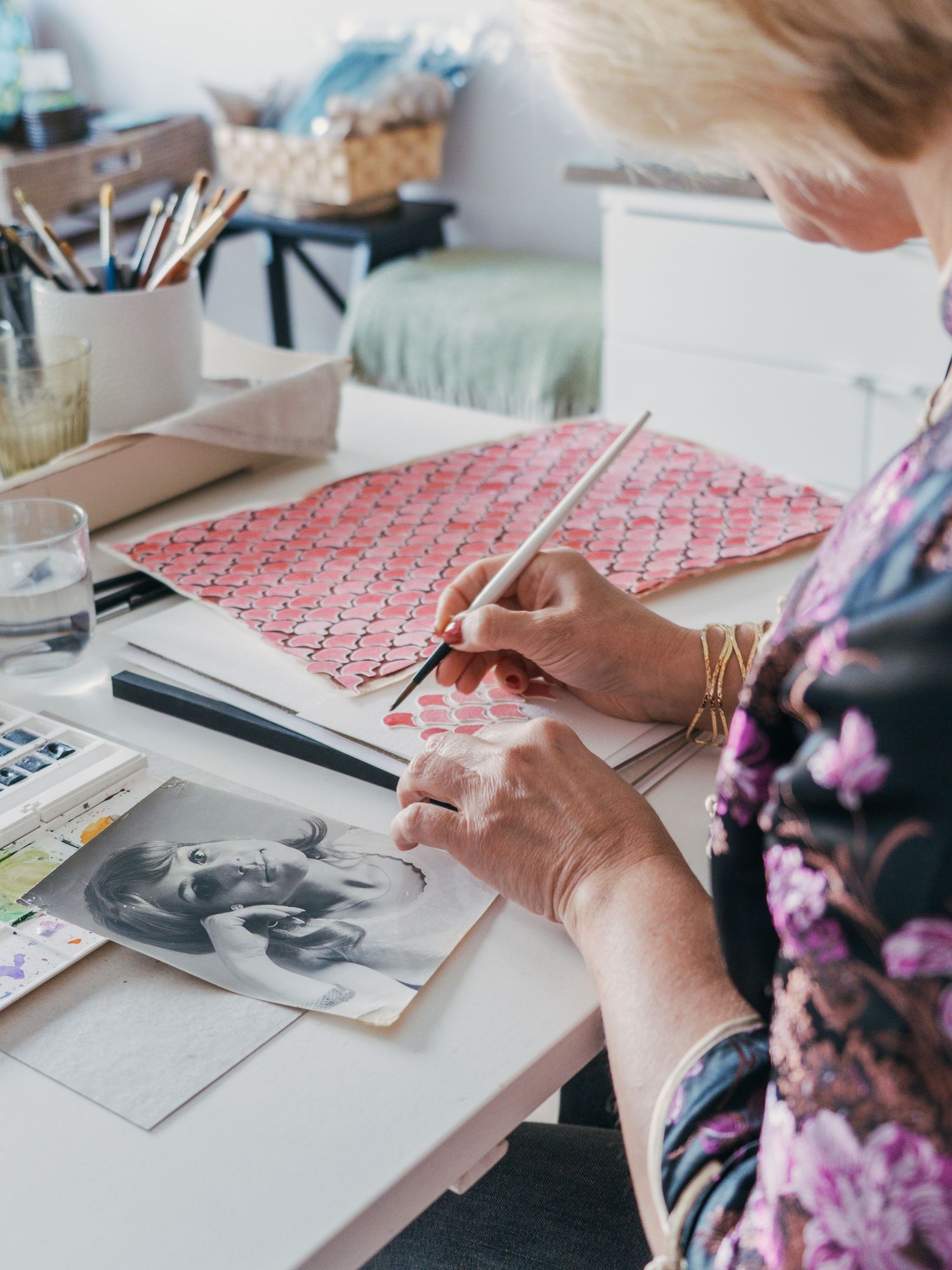 Artist working at desk with pink patterned paper and art supplies while creating detailed illustrations.