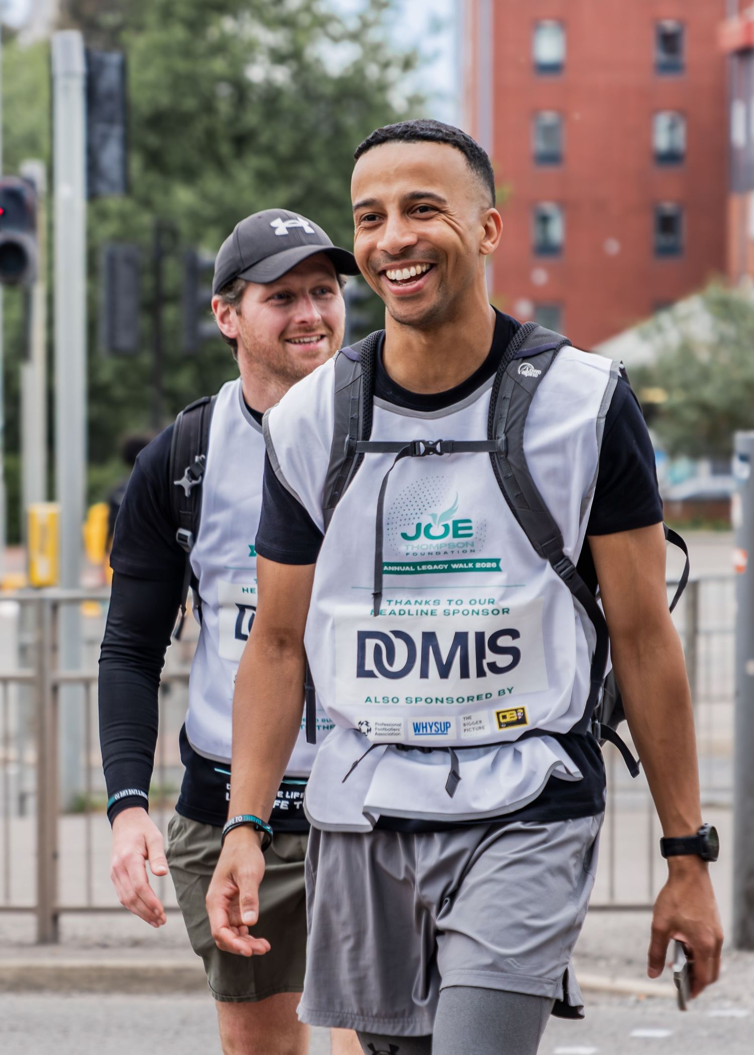 Two smiling men wearing DOMIS sponsored JOE charity walk bibs walk together outdoors.