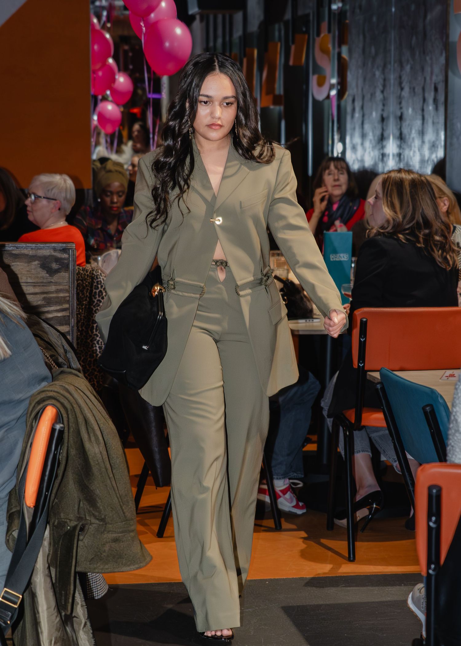 Woman in olive green suit walking through a venue decorated with pink balloons at an indoor event.