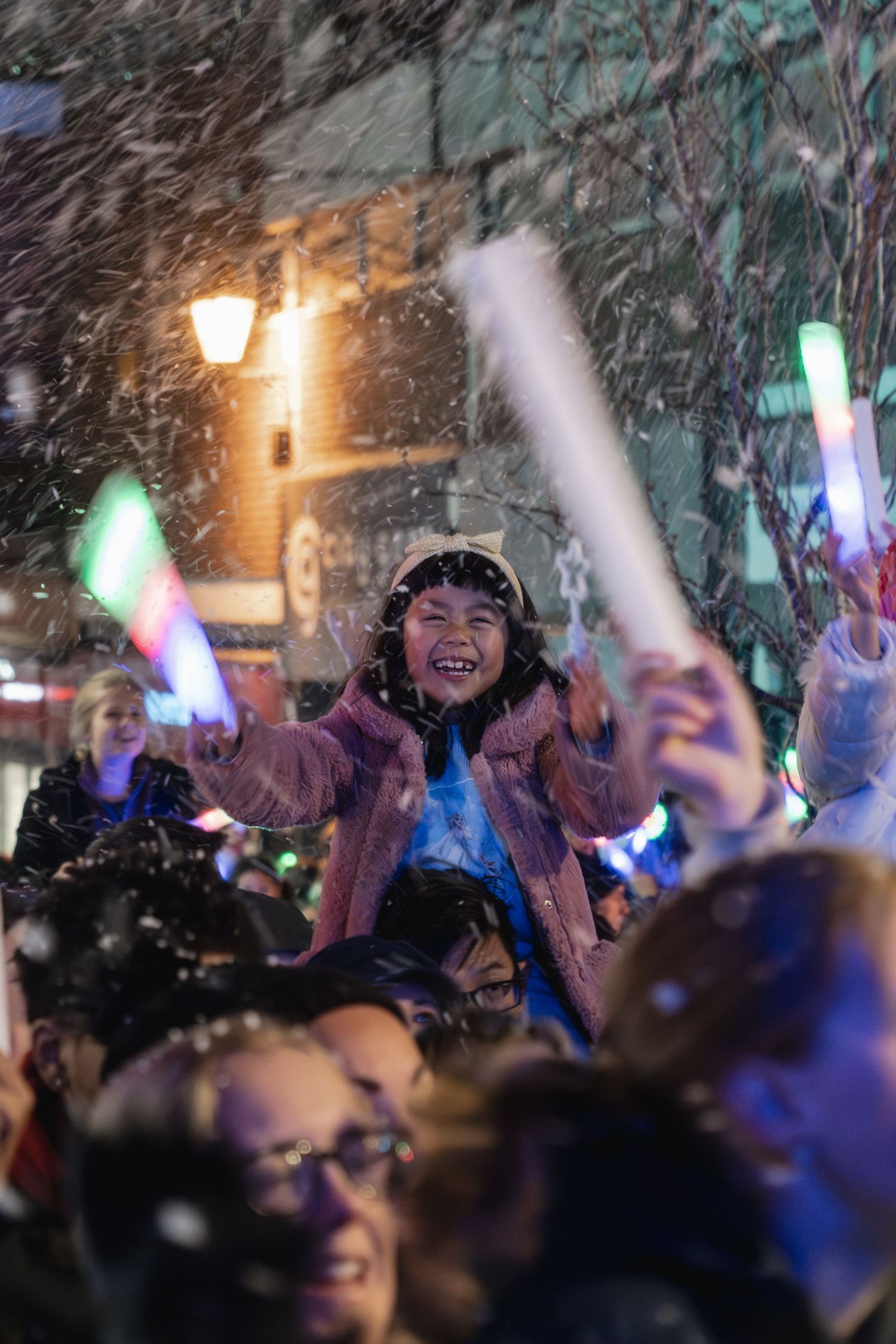 A crowd celebrates at a nighttime event with colorful light-up wands and falling snow creating a festive atmosphere.