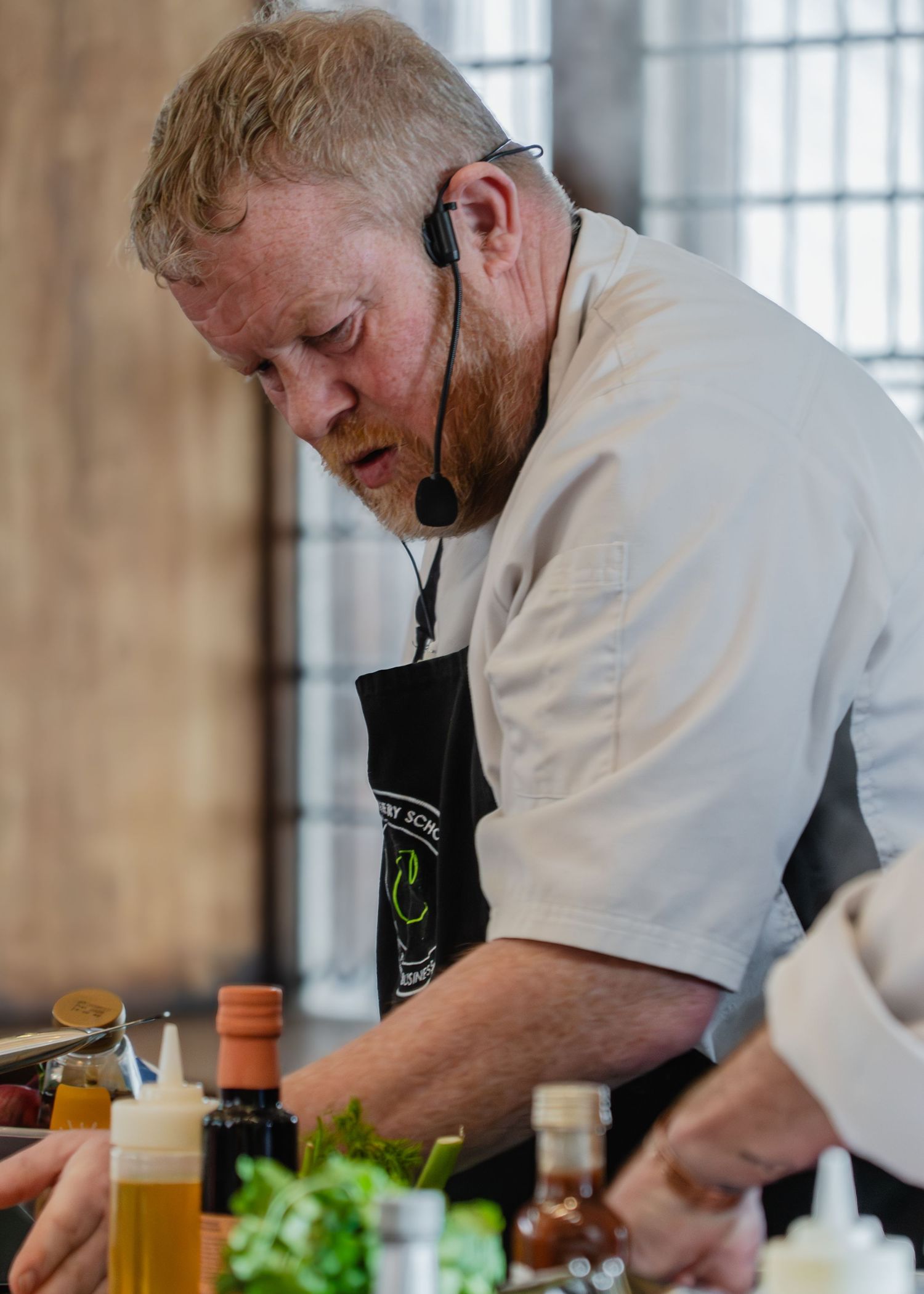 Chef in white uniform and black apron works intently at prep station with fresh ingredients and sauces.