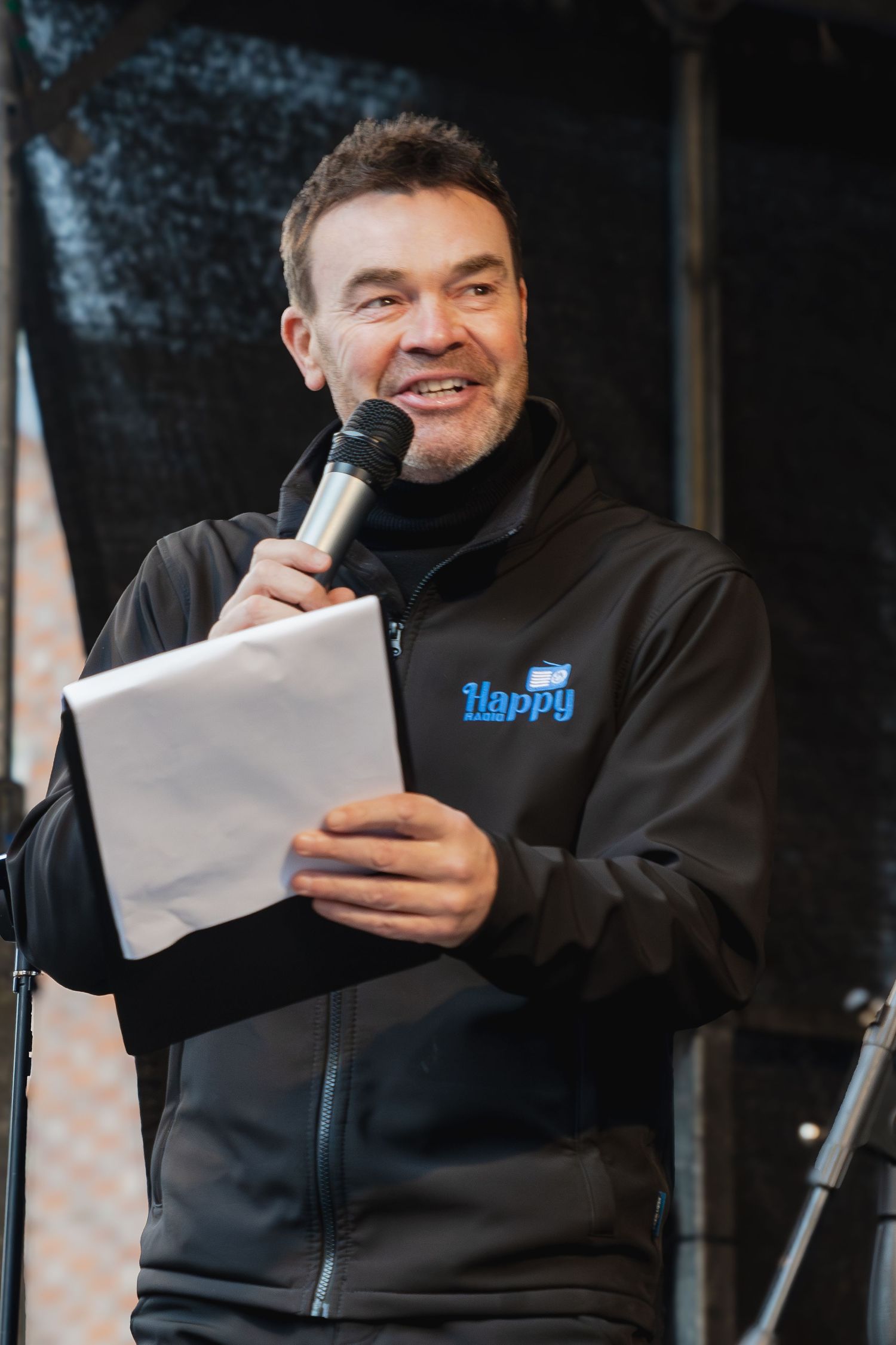 A person in a black jacket reads from papers while standing at a microphone on stage.