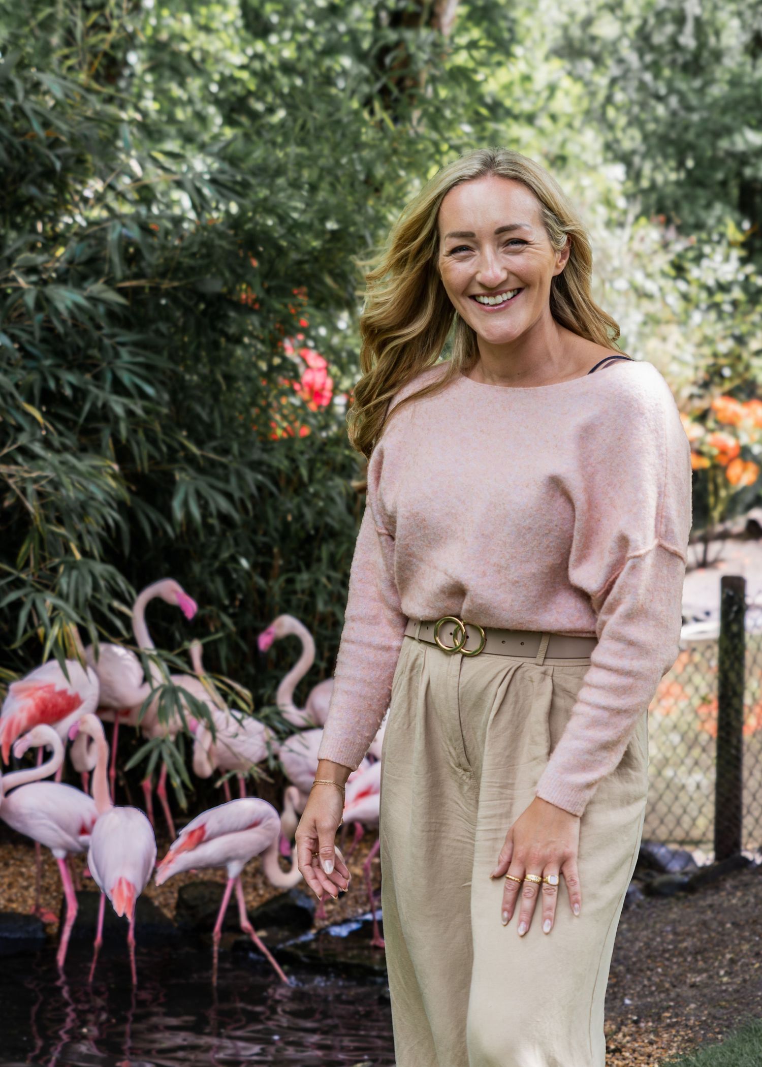 Smiling woman in pink sweater and beige trousers standing outdoors near flamingos and lush green trees.