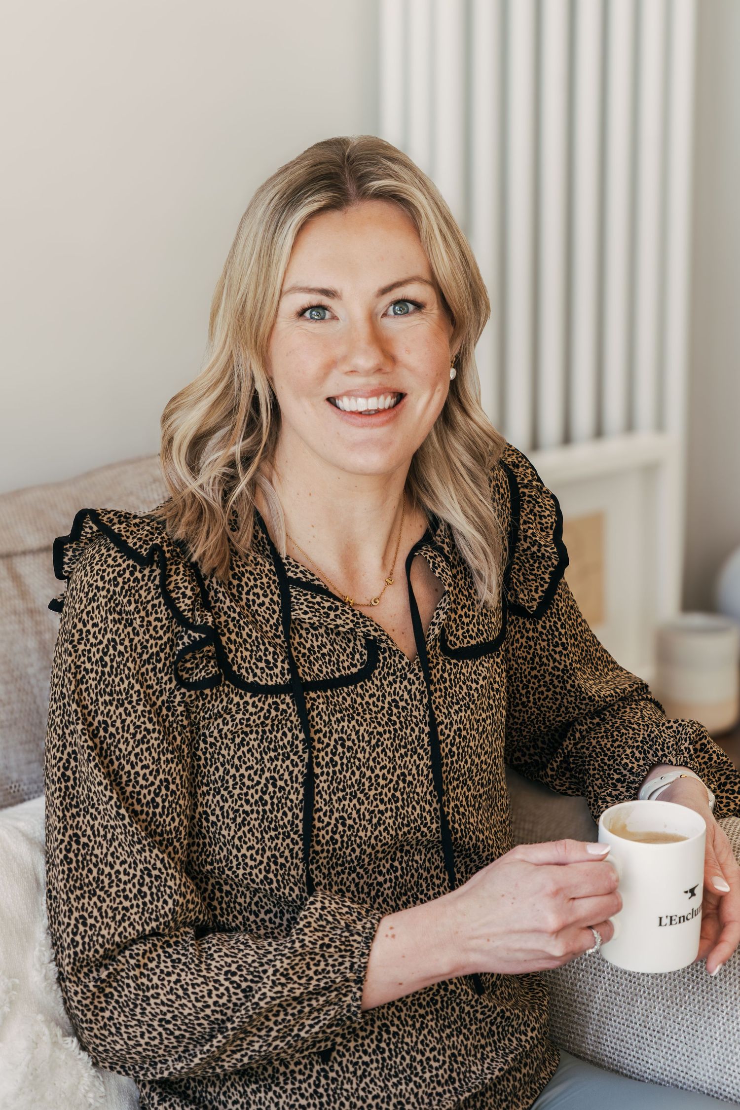 Woman in leopard print blouse smiles while holding white coffee mug in bright interior setting.