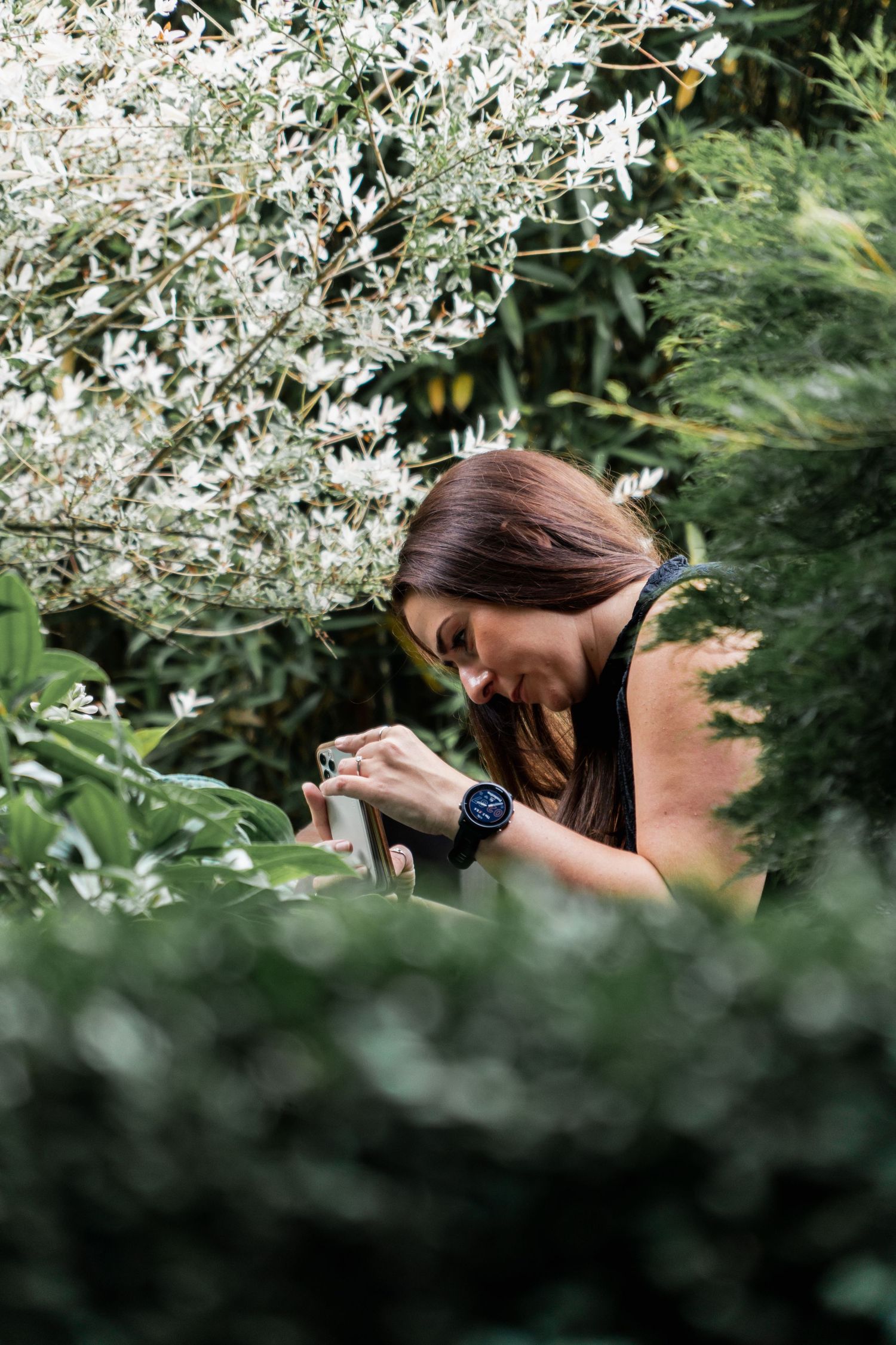 A photographer with long dark hair takes photos among delicate white spring blossoms in a natural outdoor setting.