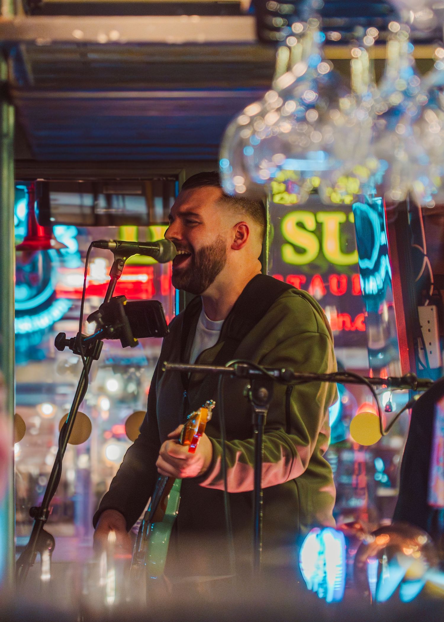 A musician performs on stage with a microphone against colorful neon lights and signage in the background.