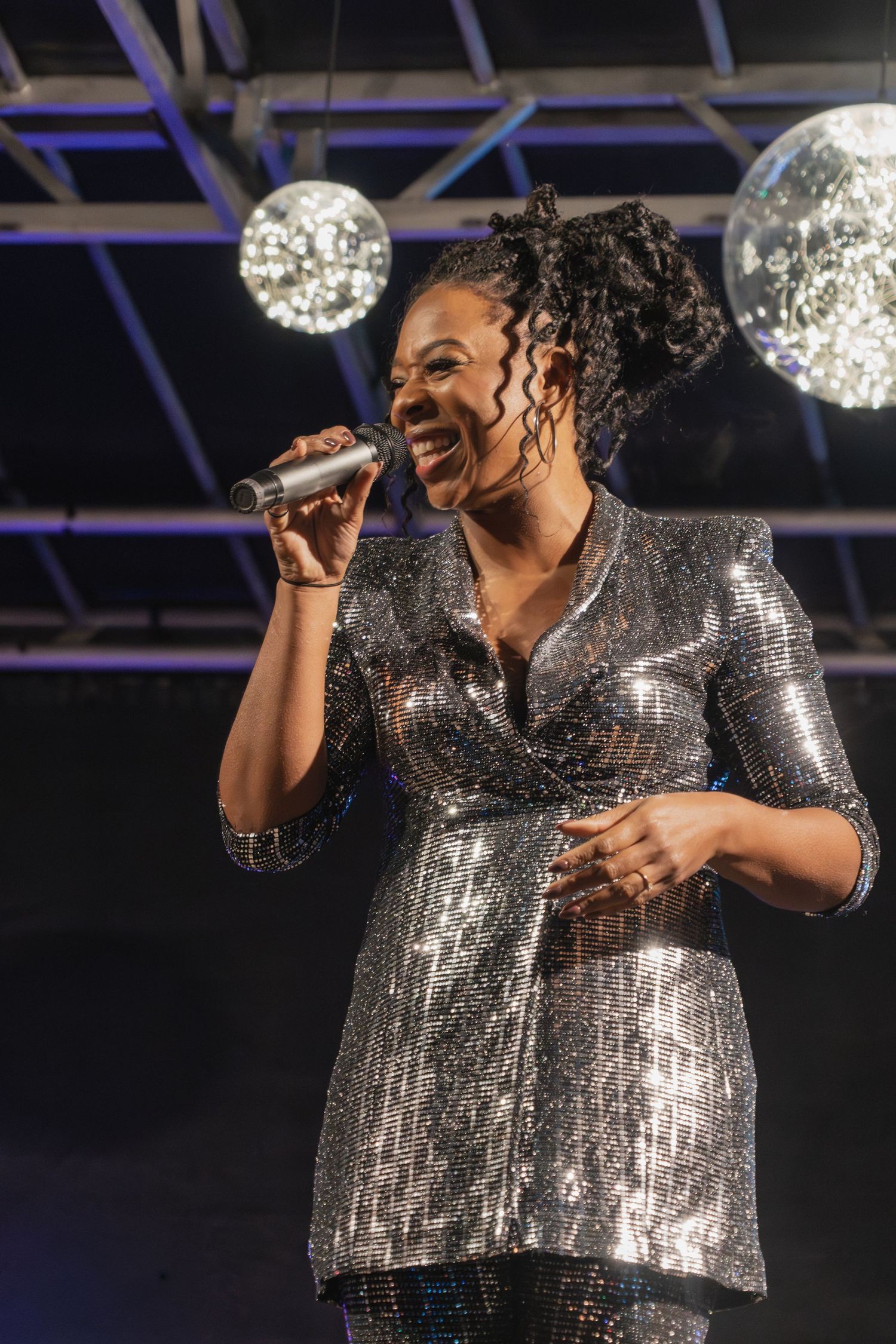 A performer in a sparkly silver dress sings passionately on stage with disco balls glowing in the background.