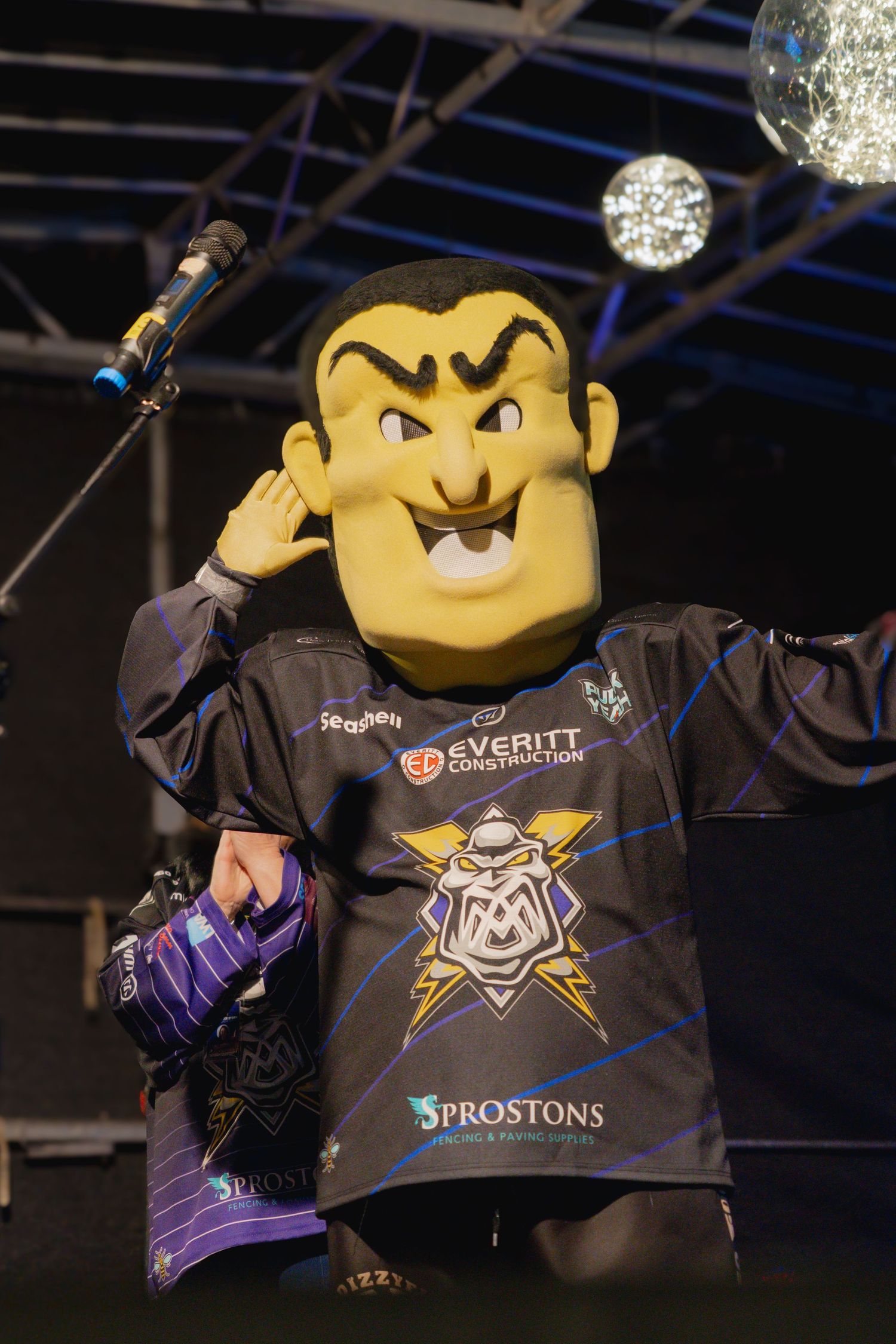A yellow cartoon mascot character wearing a black promotional t-shirt with gaming logos displayed at an event.