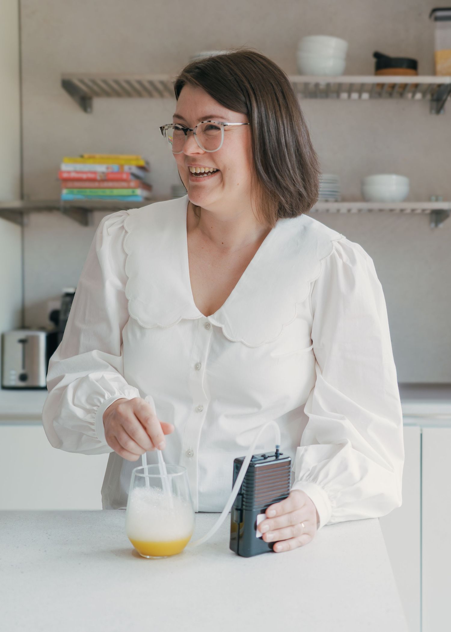 Person in white blouse smiling while using phone and drinking orange juice at bright modern kitchen counter.
